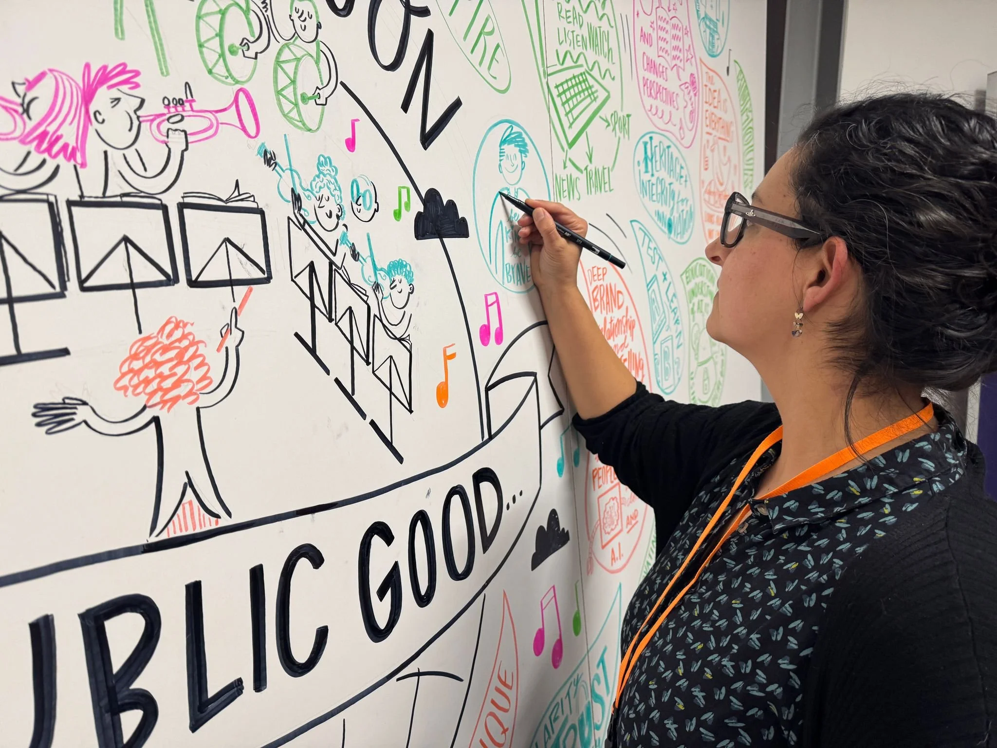 A woman with dark curly hair, glasses, and earrings draws colorful illustrations and text on a whiteboard. The illustrations include musical notes, people, and various designs. The woman is adding to a large colorful poster or mural with the phrase "