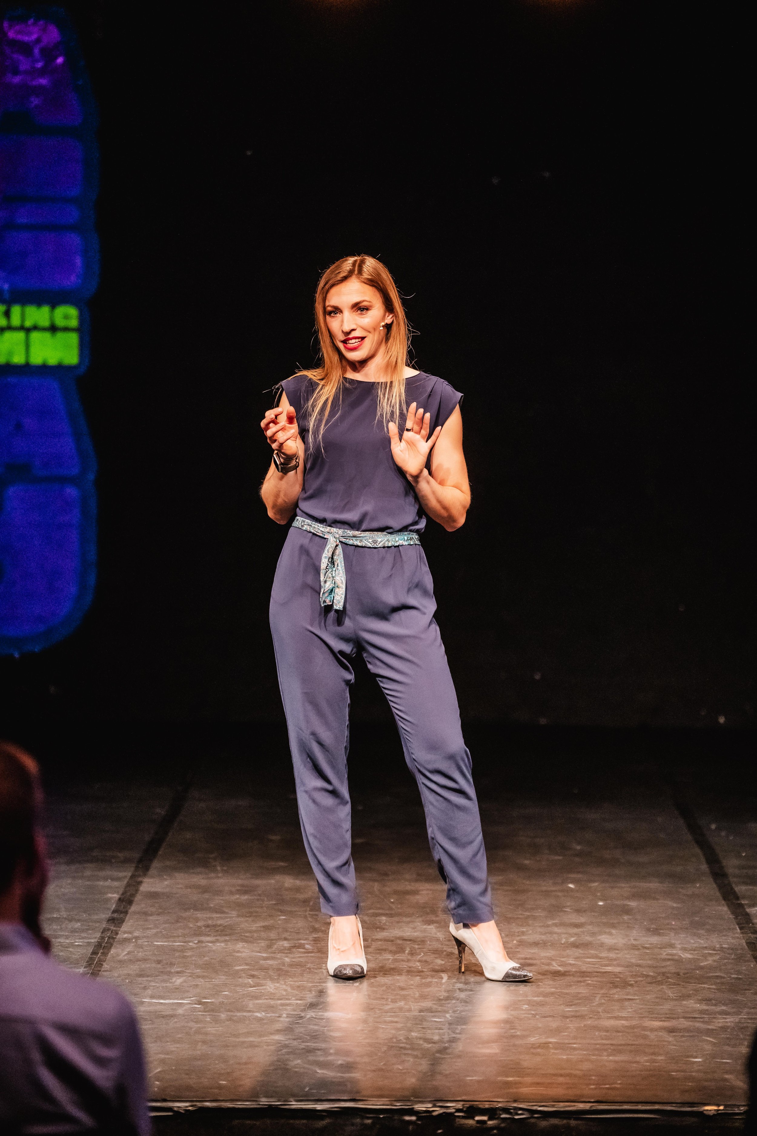 Frau spricht bei einer Präsentation auf der Bühne, schwarze Hintergrund, Trägerin eines blauen Outfits, beige Schuhe, Kopfhörer mit Mikrofon
