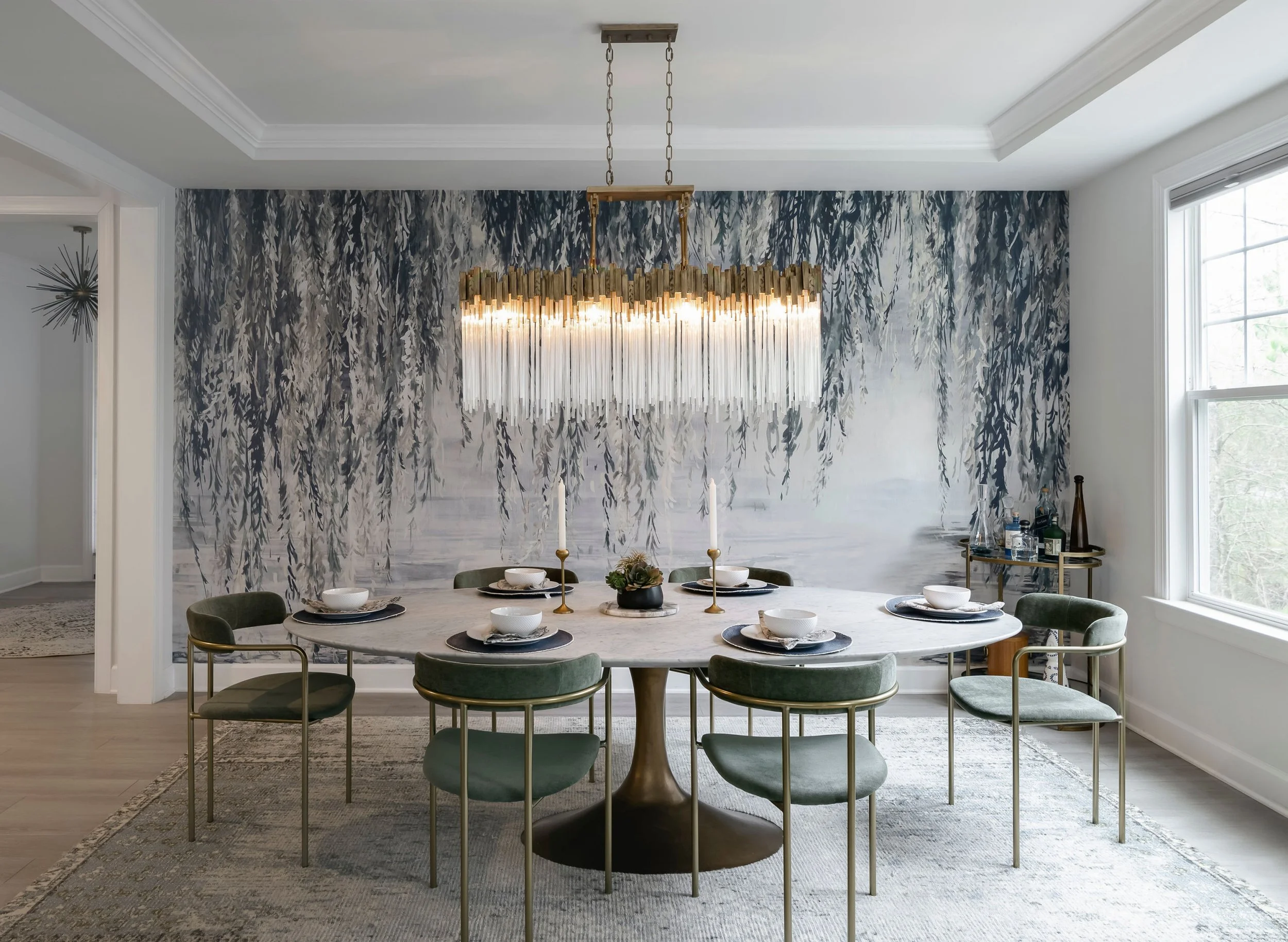 Elegant dining room with a round marble table set for six, surrounded by green velvet chairs with gold accents, a modern chandelier overhead, and a large landscape mural of a hanging willow tree on the wall.