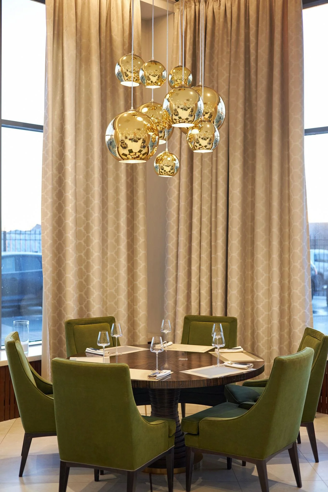 Elegant dining area with a round wooden table, six green upholstered chairs, a modern chandelier with multiple reflective gold and black orbs, beige patterned curtains, and large windows.