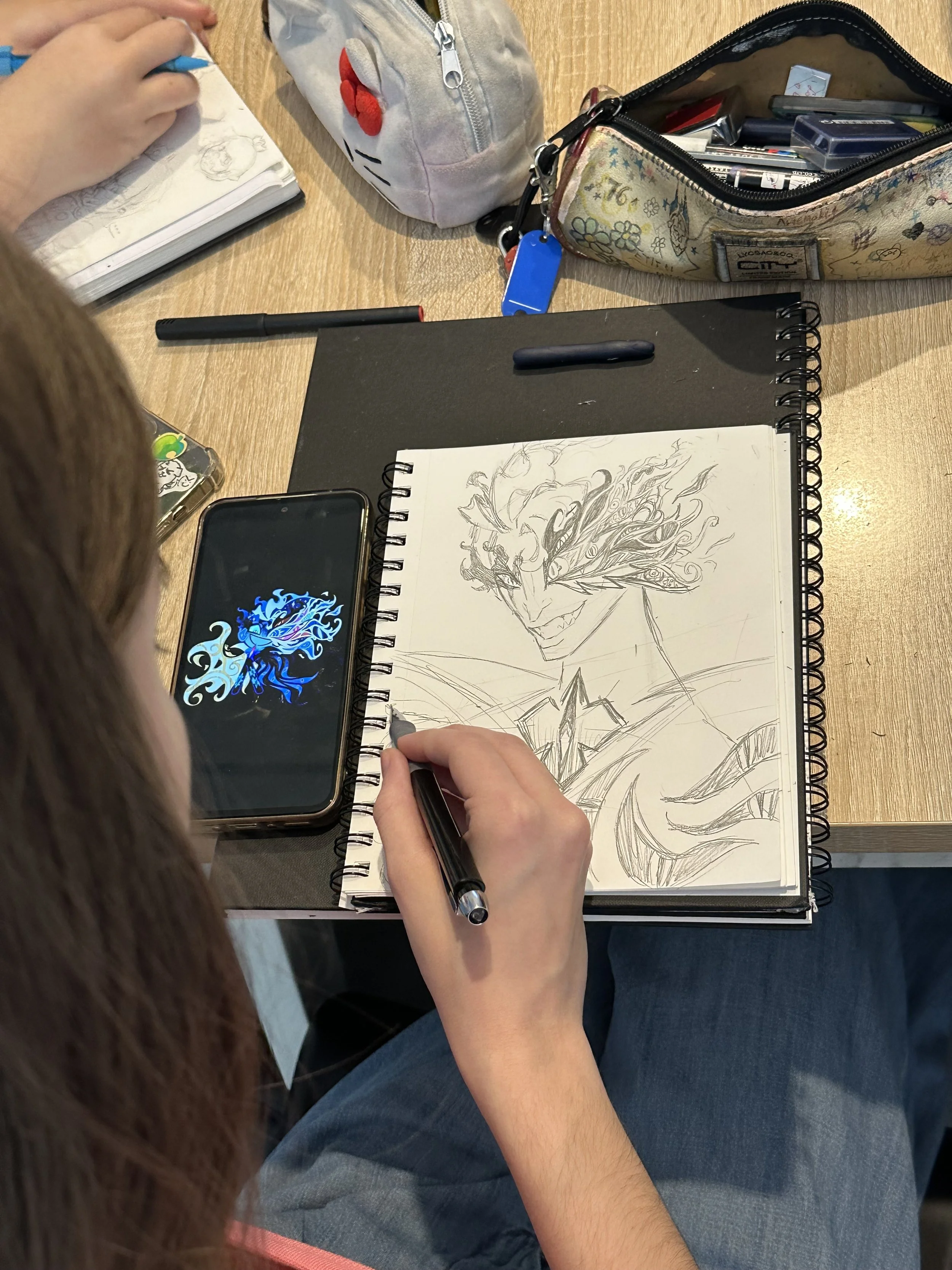 A person is drawing a detailed portrait of a smiling man with a headscarf and earrings in a sketchbook using a black pen. The person's right hand holds the pen, and the left hand adjusts the sketchbook. The workspace includes a black notebook, a black marker, a smartphone displaying a colorful dragon logo, a pencil case with art supplies, and partially visible notebooks or sketchbooks. The person is wearing a gray shirt and blue jeans, sitting on a chair at a wooden table.