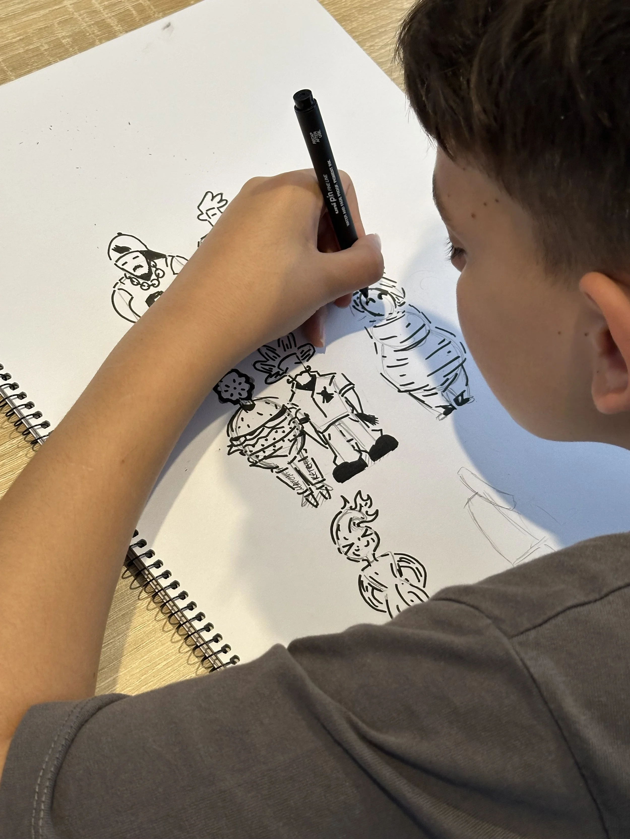 A person is coloring or drawing on a large white sheet of paper on a notebook with a black marker. The drawing includes several cartoon-like characters, such as a princess, a knight, and a girl with pigtails.