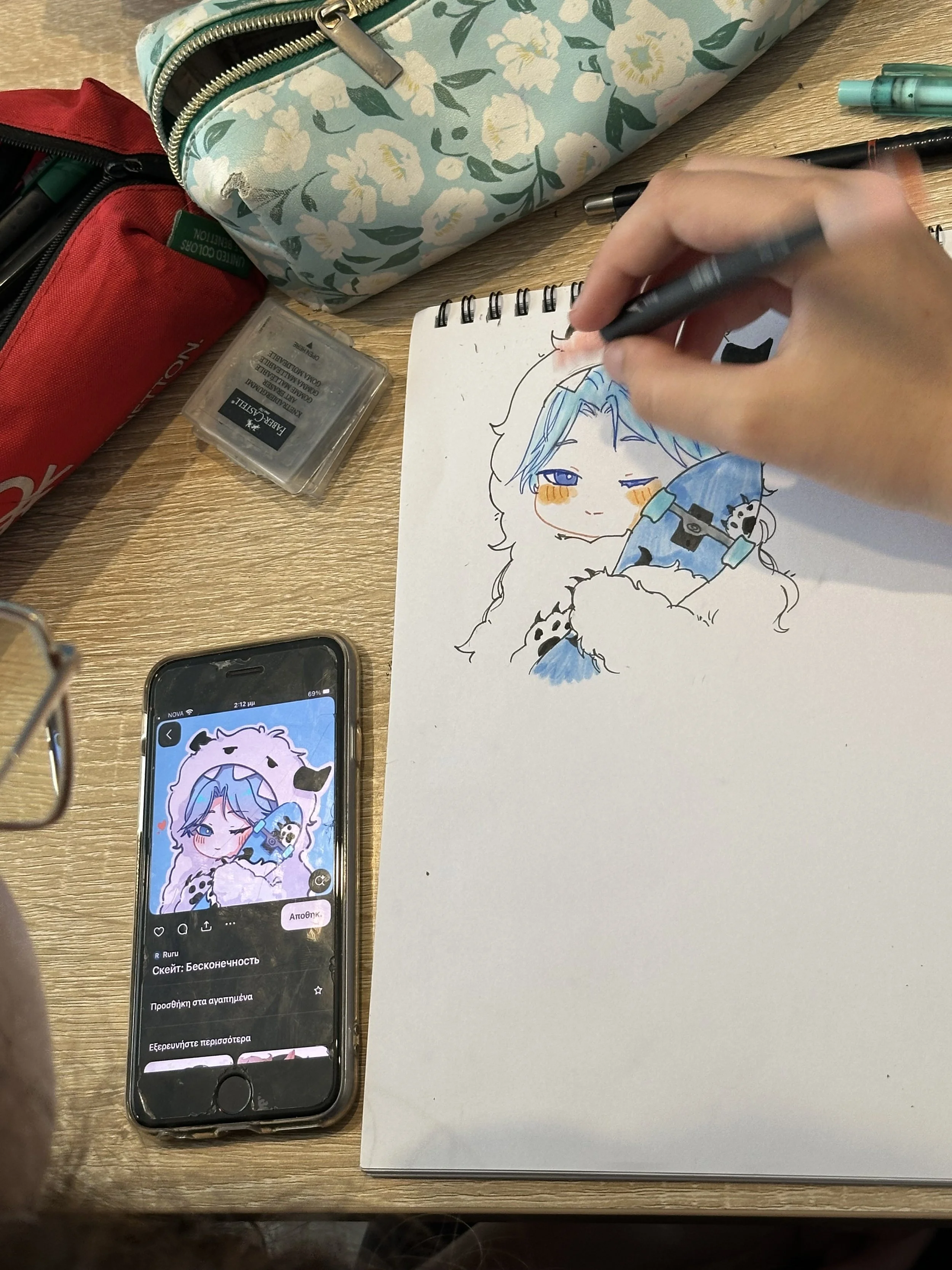 A person is drawing a colorful anime-style character on a spiral sketchpad with a black pen, along with stationery items including a floral-patterned pencil case, a red pencil case, markers, and a smartphone displaying an illustration of the same character.