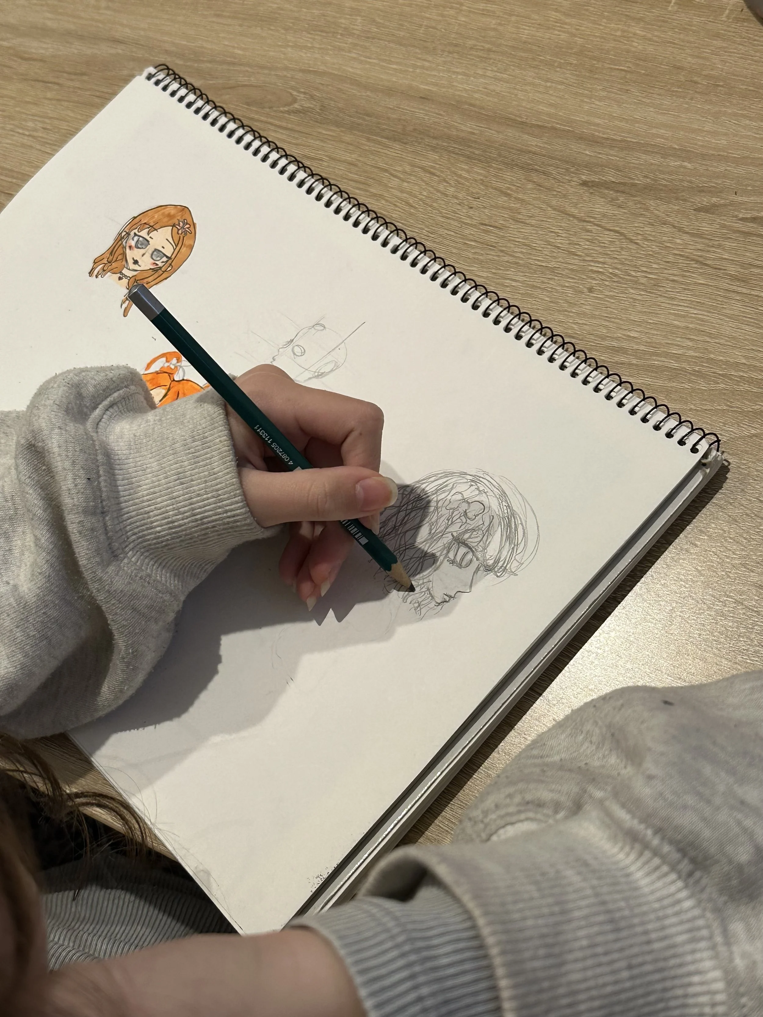 Drawing of a girl with orange hair, flower accessory, big eyes, and dark lips in a sketchbook, with other sketches visible.