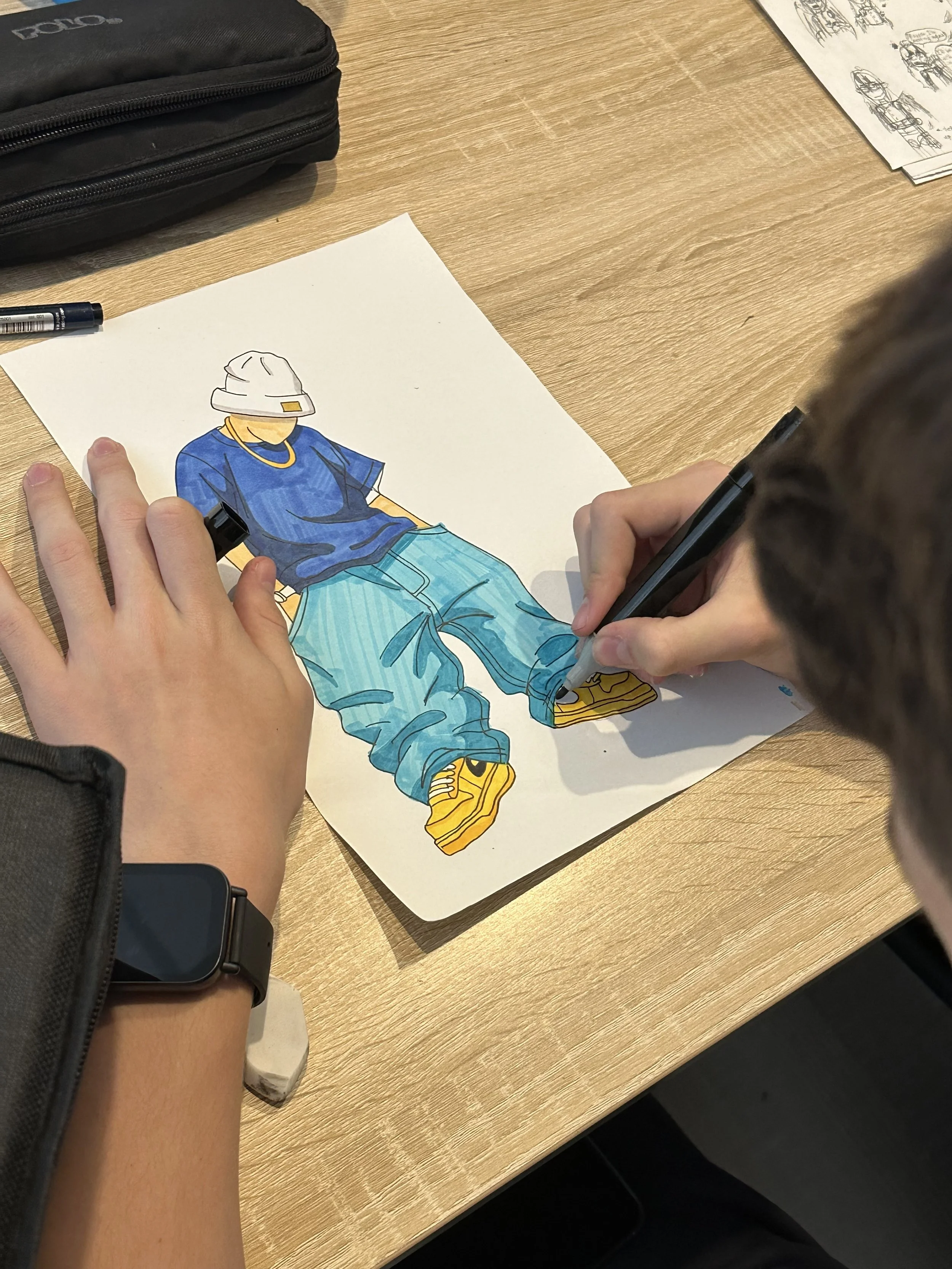 Person drawing and coloring a fashion illustration of a model wearing a blue shirt, light blue pants, yellow sneakers, and a white hat, on a white sheet of paper on a wooden table.