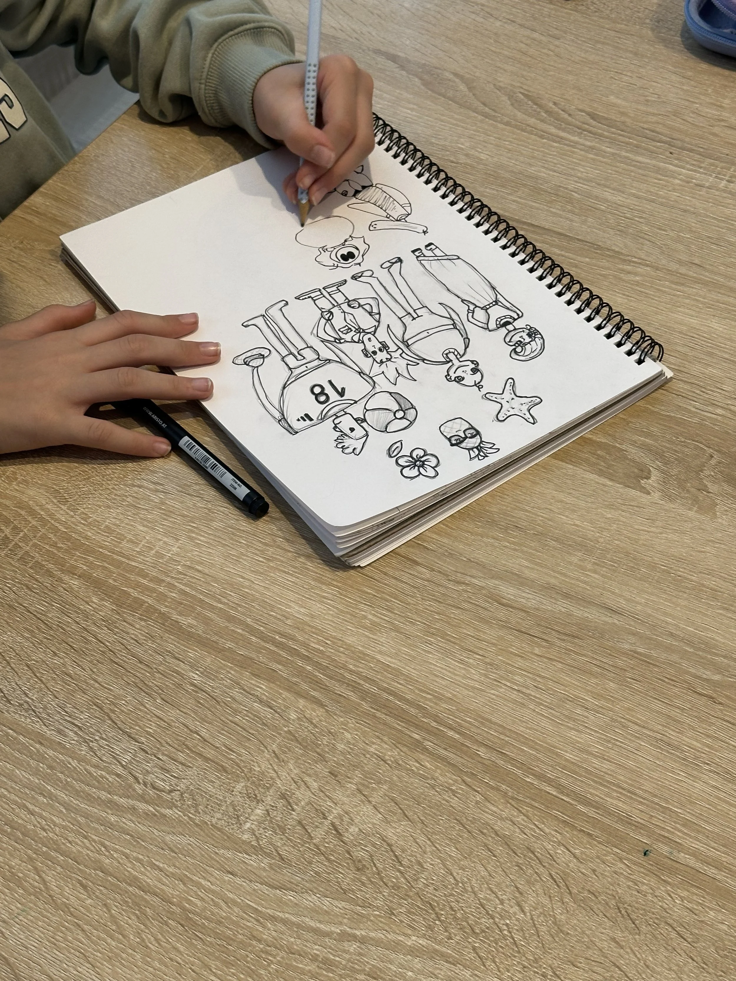 A person is coloring a drawing of various cartoon characters, including a dinosaur, a starfish, a flower, and animals, in a spiral-bound sketchbook on a wooden table.