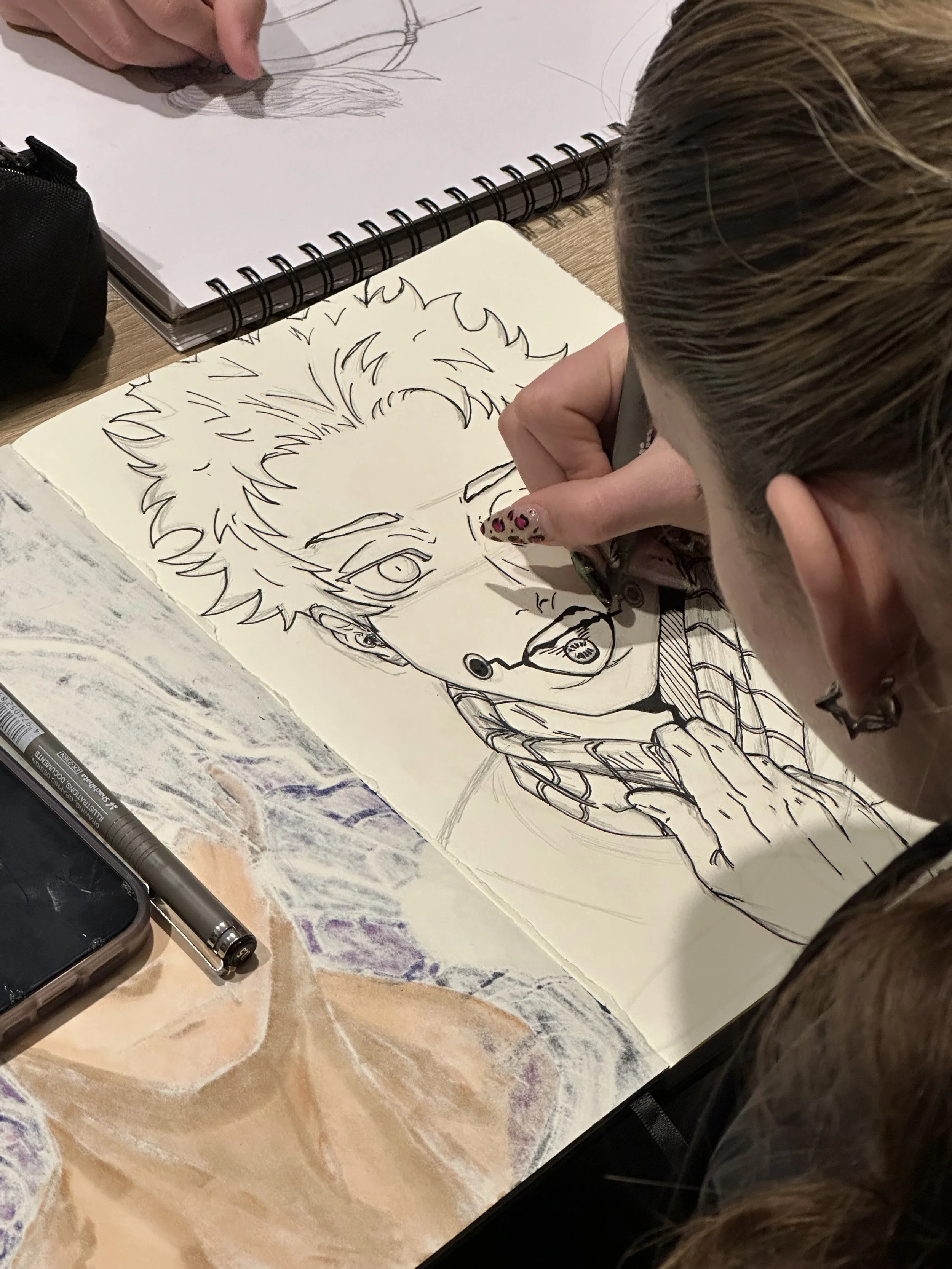 A person is drawing a detailed portrait of a person with curly hair on a sheet of paper, using a pen. The drawing features expressive facial details. The person's hand is visible, and they have painted nails with a leopard print design.