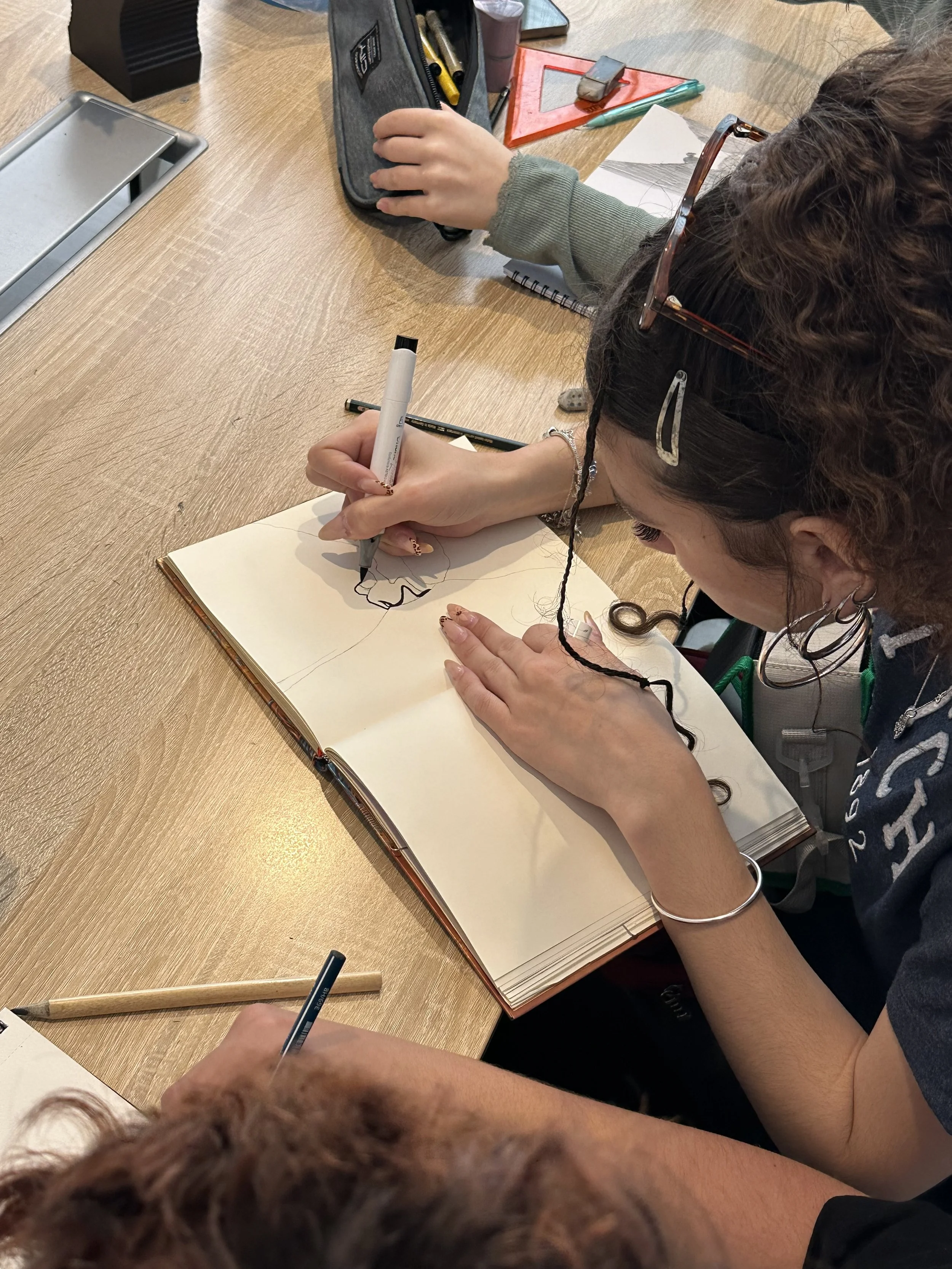 A person is drawing or writing in a sketchbook at a wooden tabletop, with another person beside them. Various stationery items such as pens, markers, a ruler, and a pencil case are on the table.