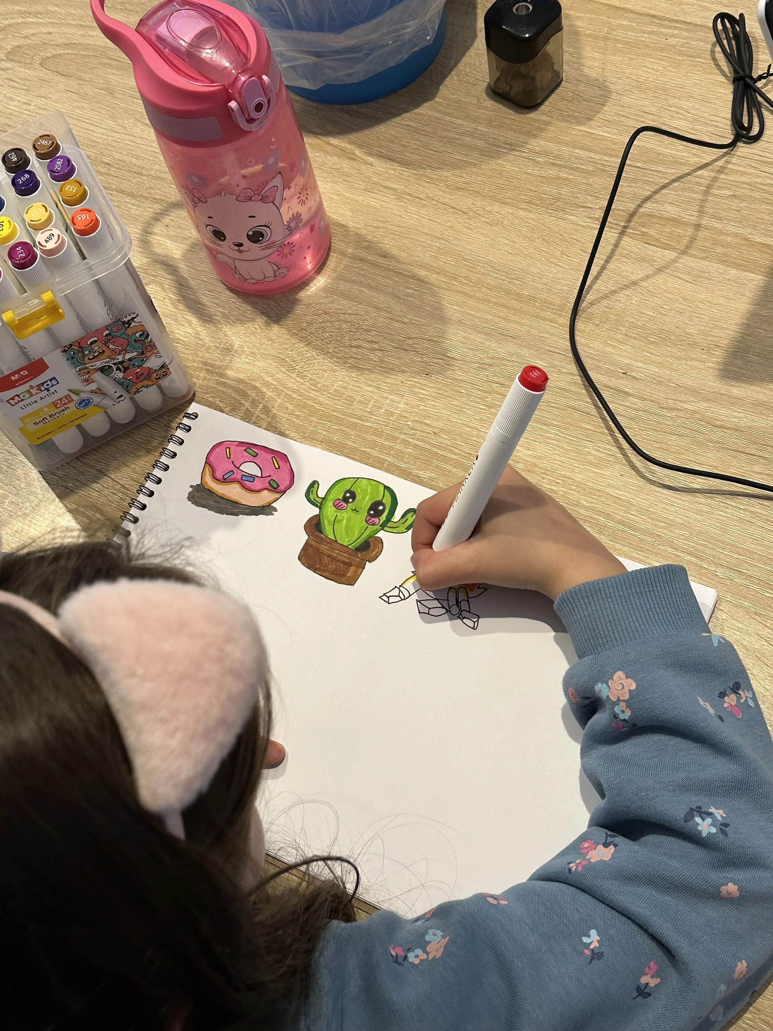 A child drawing a colorful picture of a cactus with a pink donut on top on a sketchpad, with a pink water bottle and colored markers nearby on a wooden table.