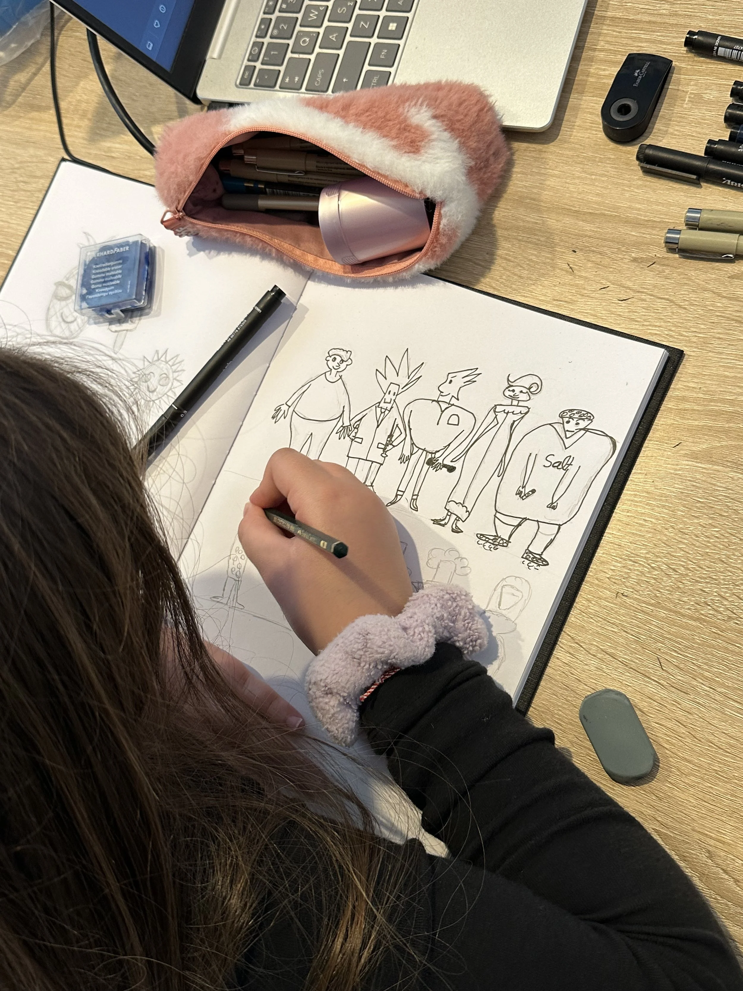 Drawing of cartoon characters on a sketchbook with a person using a black pen, surrounded by markers, a laptop, and stationery on a wooden desk.