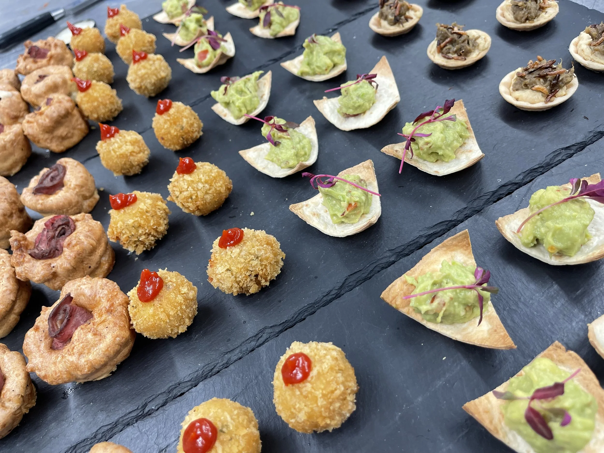 Assorted appetizer bites on a black slate platter, including fried croquettes topped with red sauce, stuffed pastry shells with avocado and microgreens, and small fried dough balls with sauce.