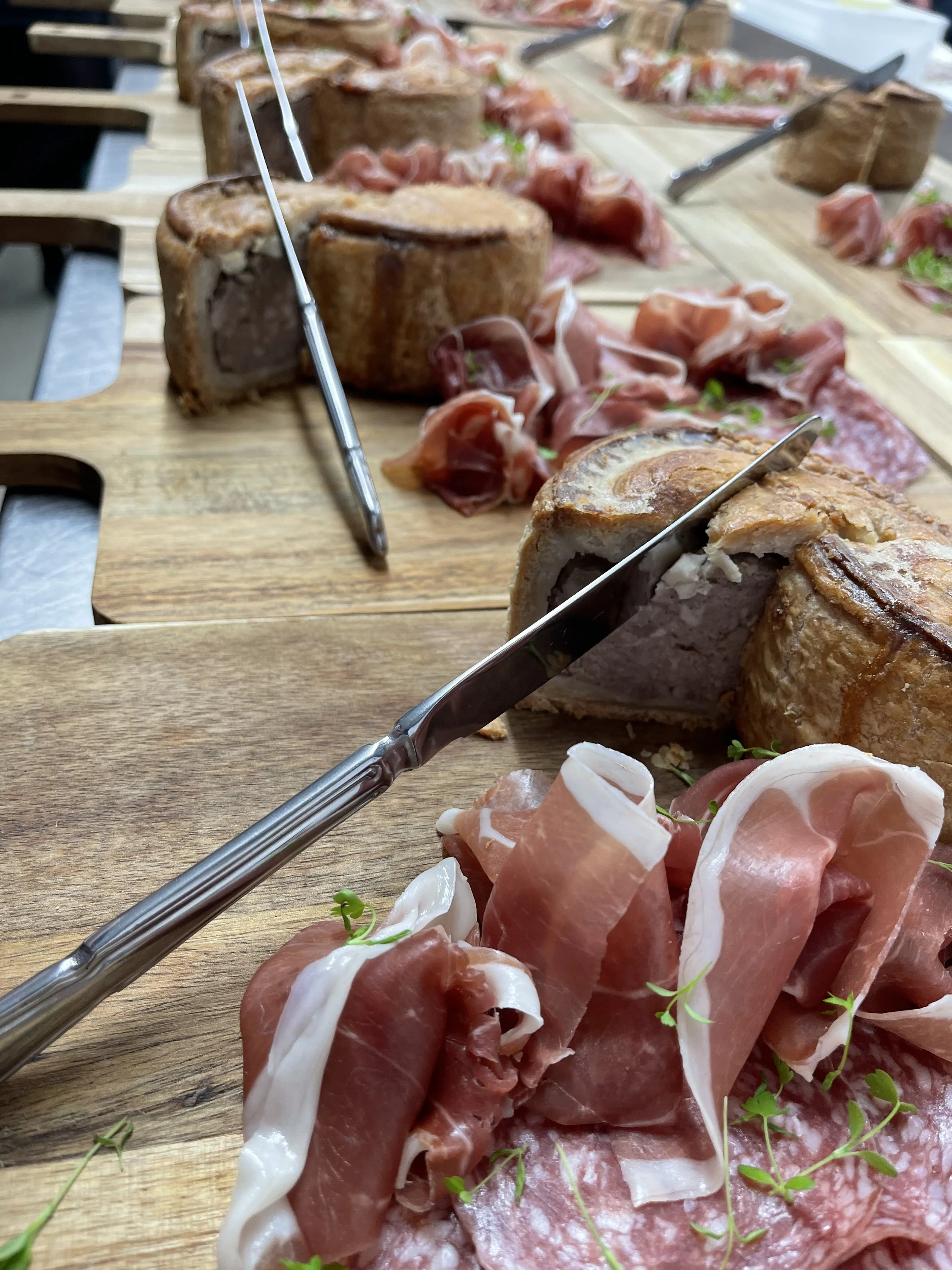 A wooden platter with slices of prosciutto and cured meats, along with a piece of roasted pork topped with herbs and a knife.