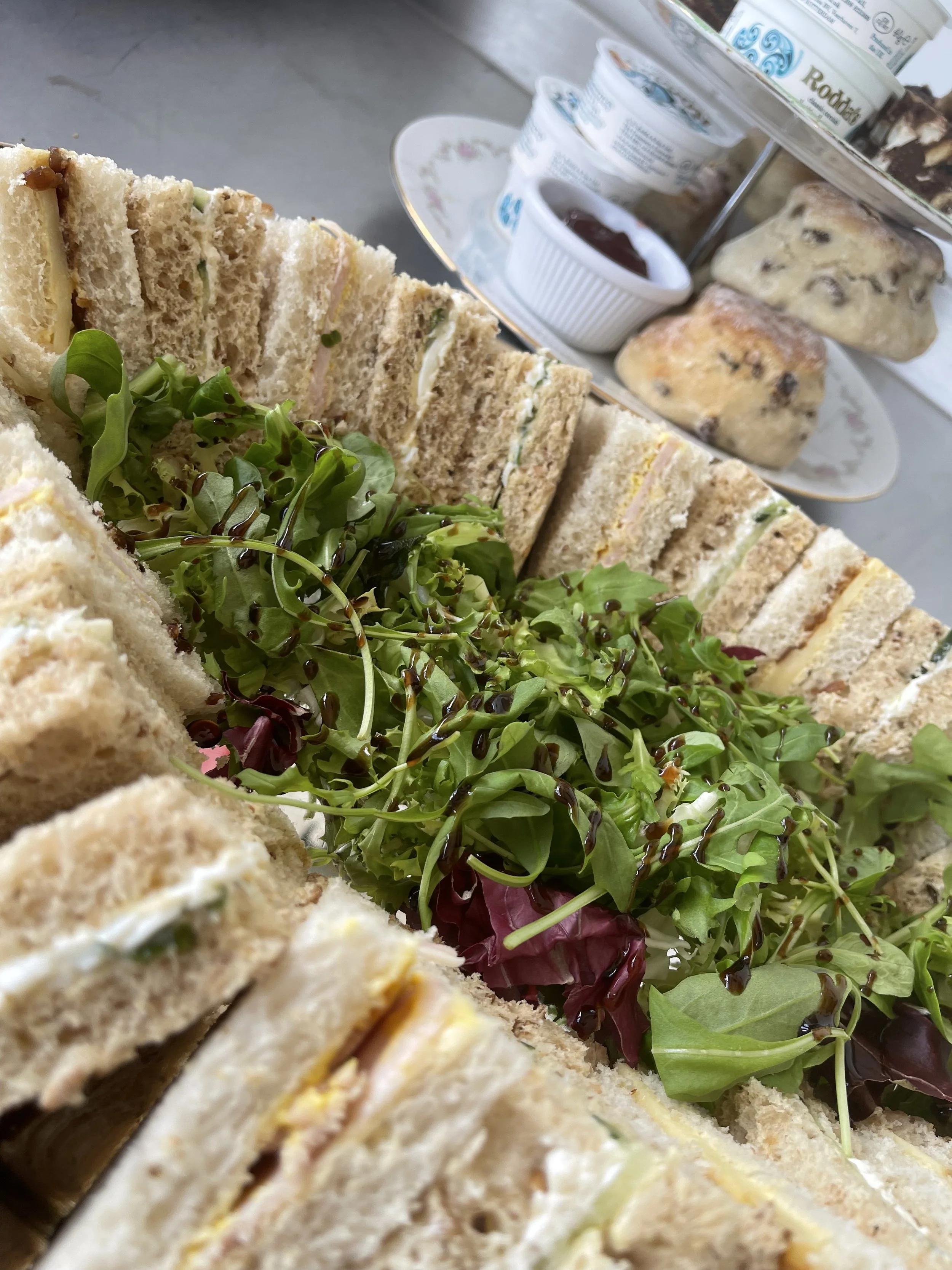 Assorted sandwiches, salad greens with balsamic glaze, and tea or coffee desserts on a multi-tiered tray on a table.