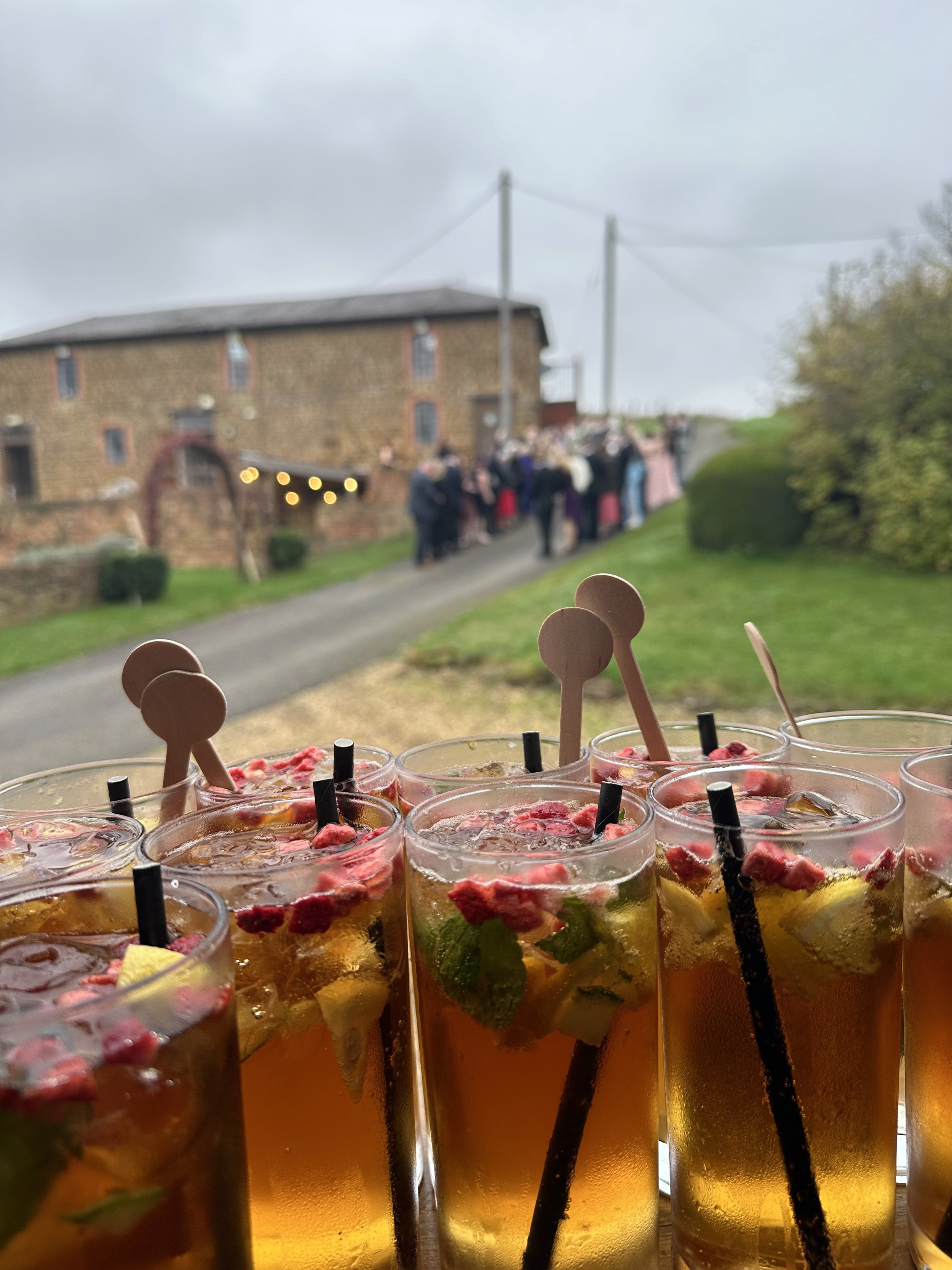 Close-up of several glasses of iced fruit cocktails with mint, lemon, and pomegranate, with cosmic black straws and wooden stir sticks, on a table outdoors during daytime at an event with a blurry crowd and rustic brick building in the background.
