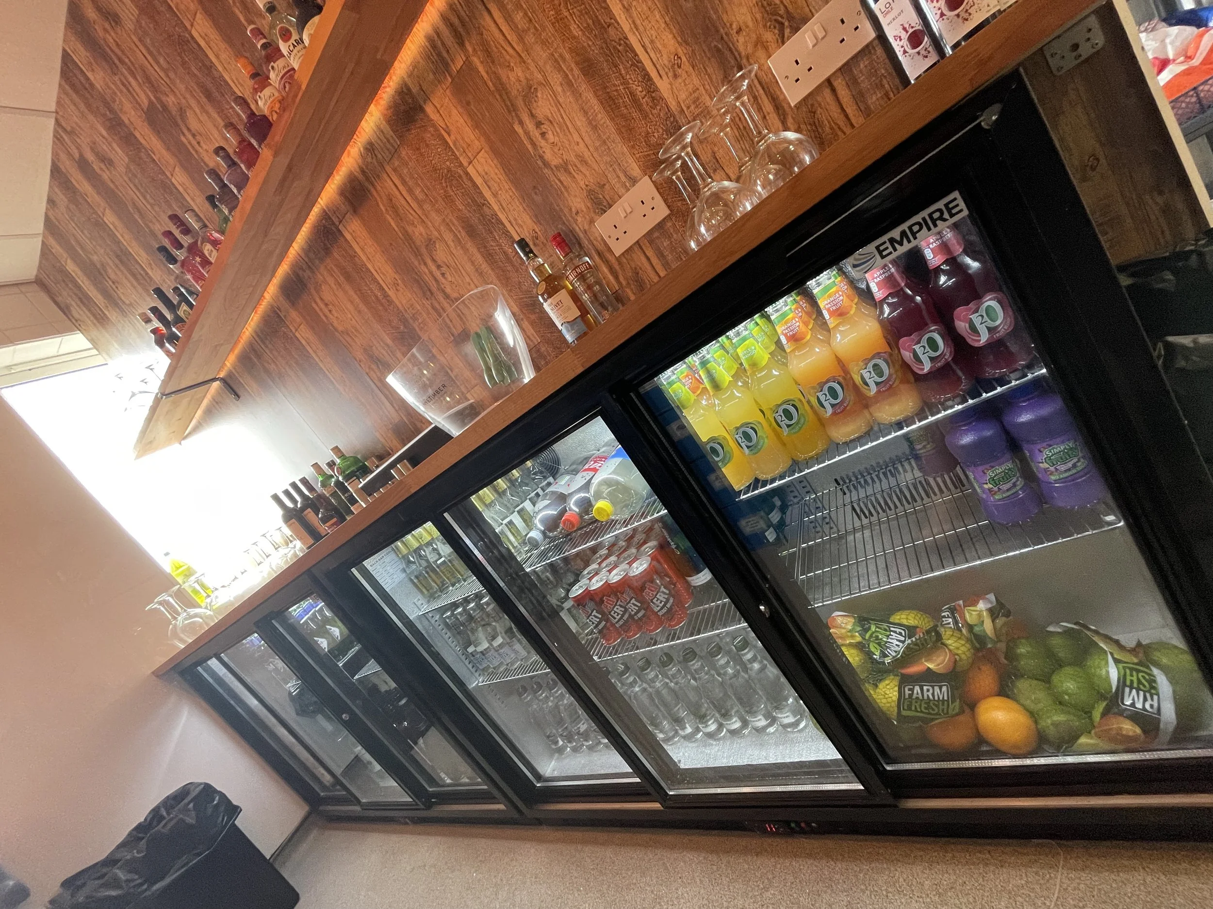 Refrigerated beverages, including orange, red, and purple drinks, along with fresh fruits like avocados, oranges, and a pineapple, are inside a glass-door fridge. The fridge is in a wooden-finished cafe or bar area with a counter above, holding bottl