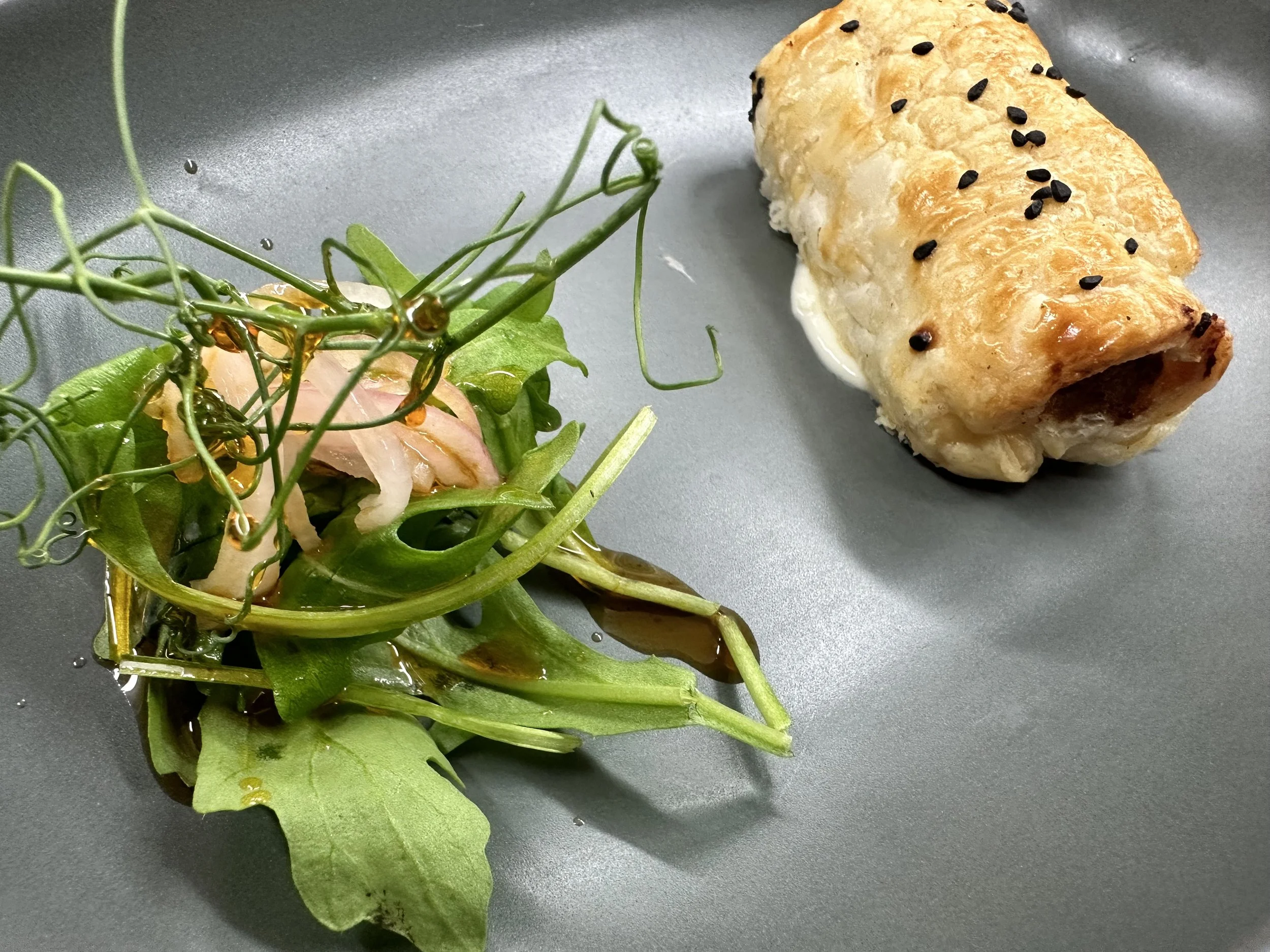 Salad with greens and thinly sliced radish next to a cooked stuffed chicken breast with black sesame seeds on top, served on a gray plate.