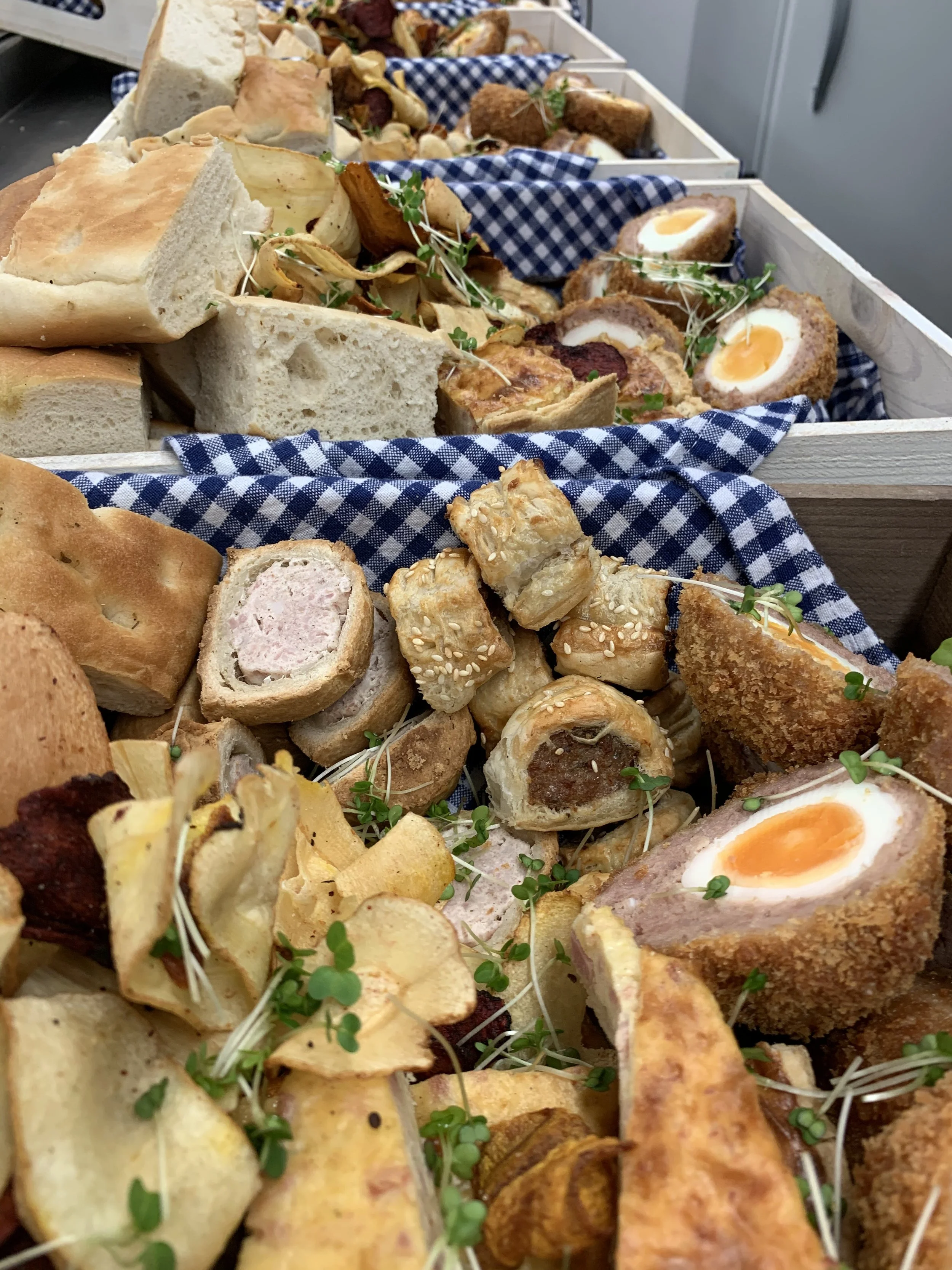 Assortment of sandwiches, bread, and fried snacks in wooden boxes lined with blue checkered cloth.