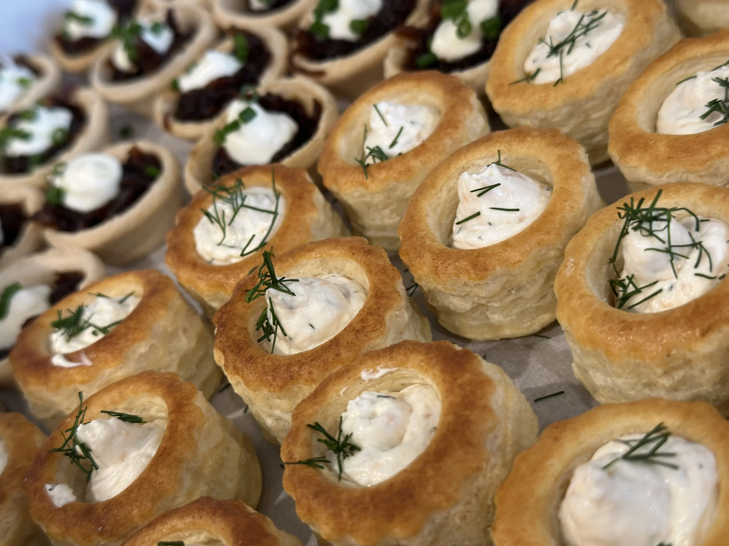 Close-up of baked savory appetizers, including cream-filled pastry cups garnished with herbs and small tart-like treats topped with a dollop of sour cream and herbs.