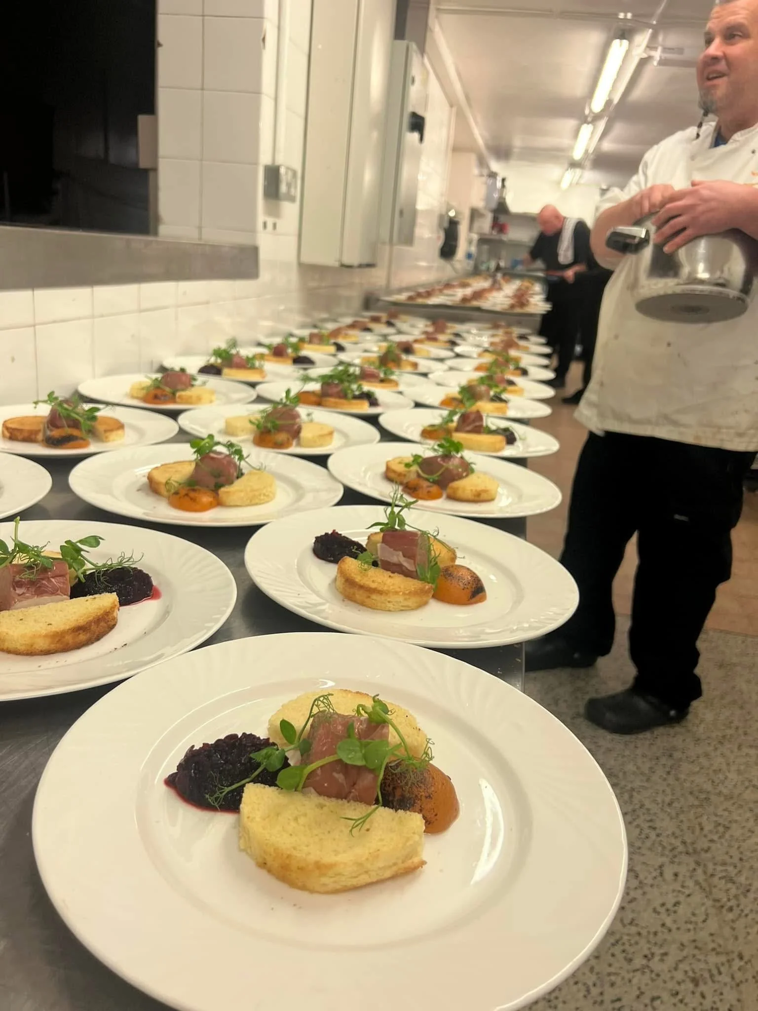 Multiple plates of gourmet appetizers on a kitchen counter, with a chef in a white uniform preparing more dishes in the background.