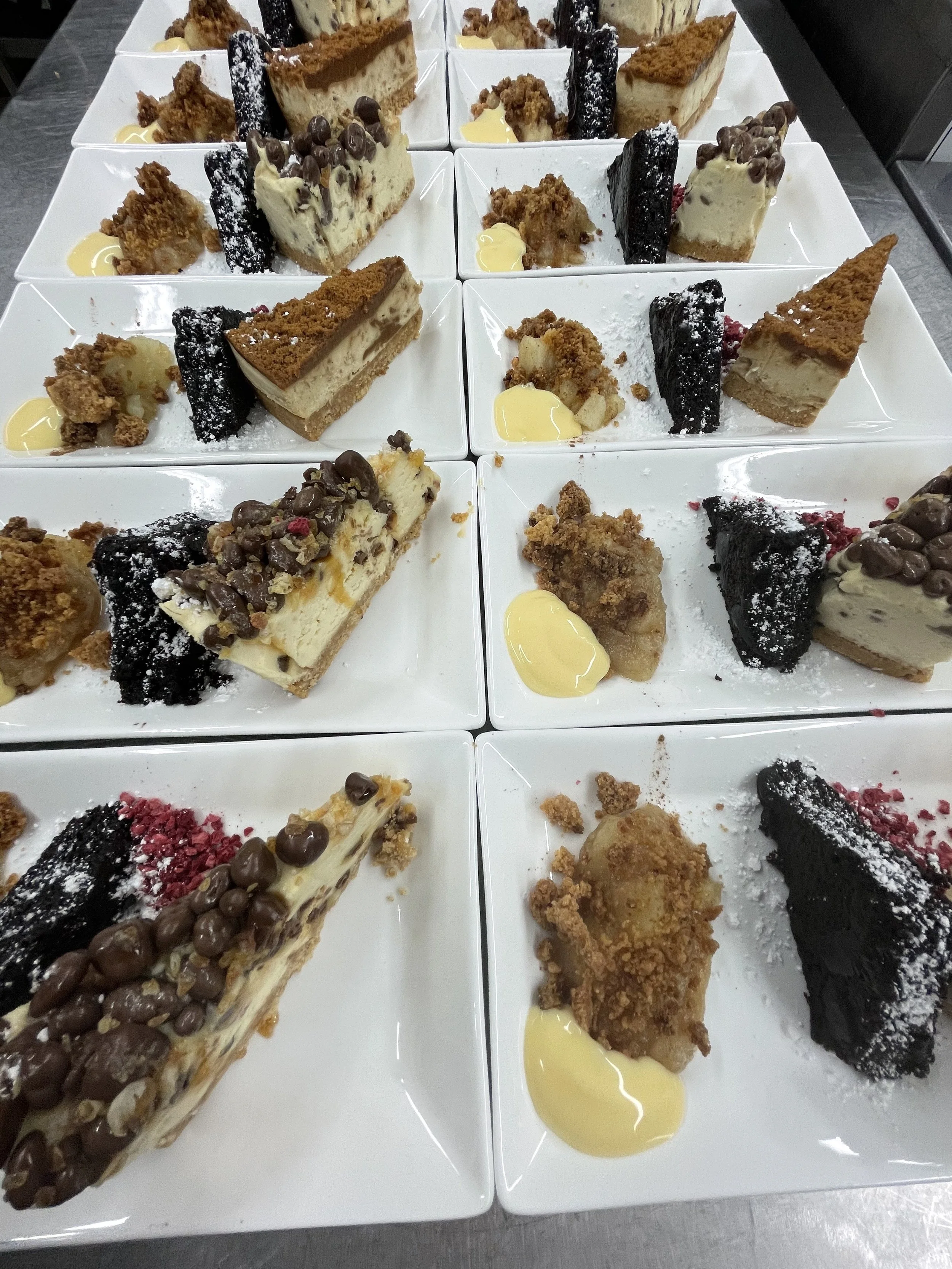 Nine white plates arranged in three rows, each containing an assortment of individual dessert slices, including cheesecakes topped with chocolate chips and cookie crumbles, layers of creamy fillings with cookie crusts, and slices of chocolate or cook