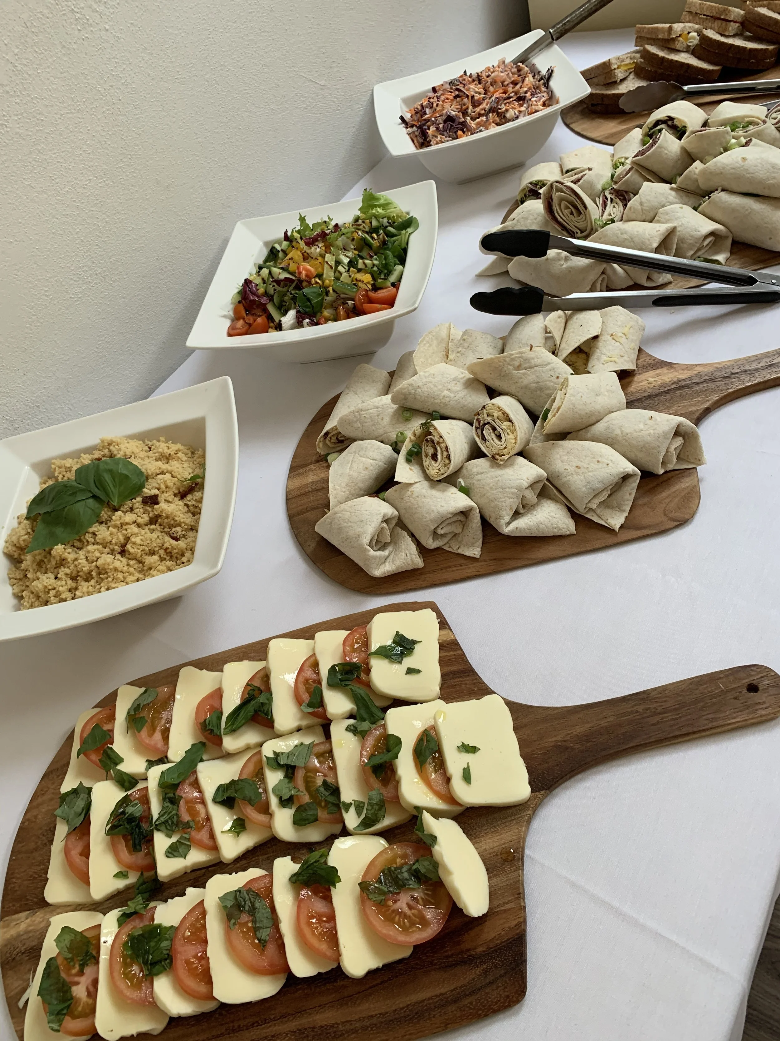 A buffet table with various appetizers including tomato and cheese platter, wraps, a salad, a grain dish, and a relish or shredded meat, all arranged on wooden serving boards and white bowls.