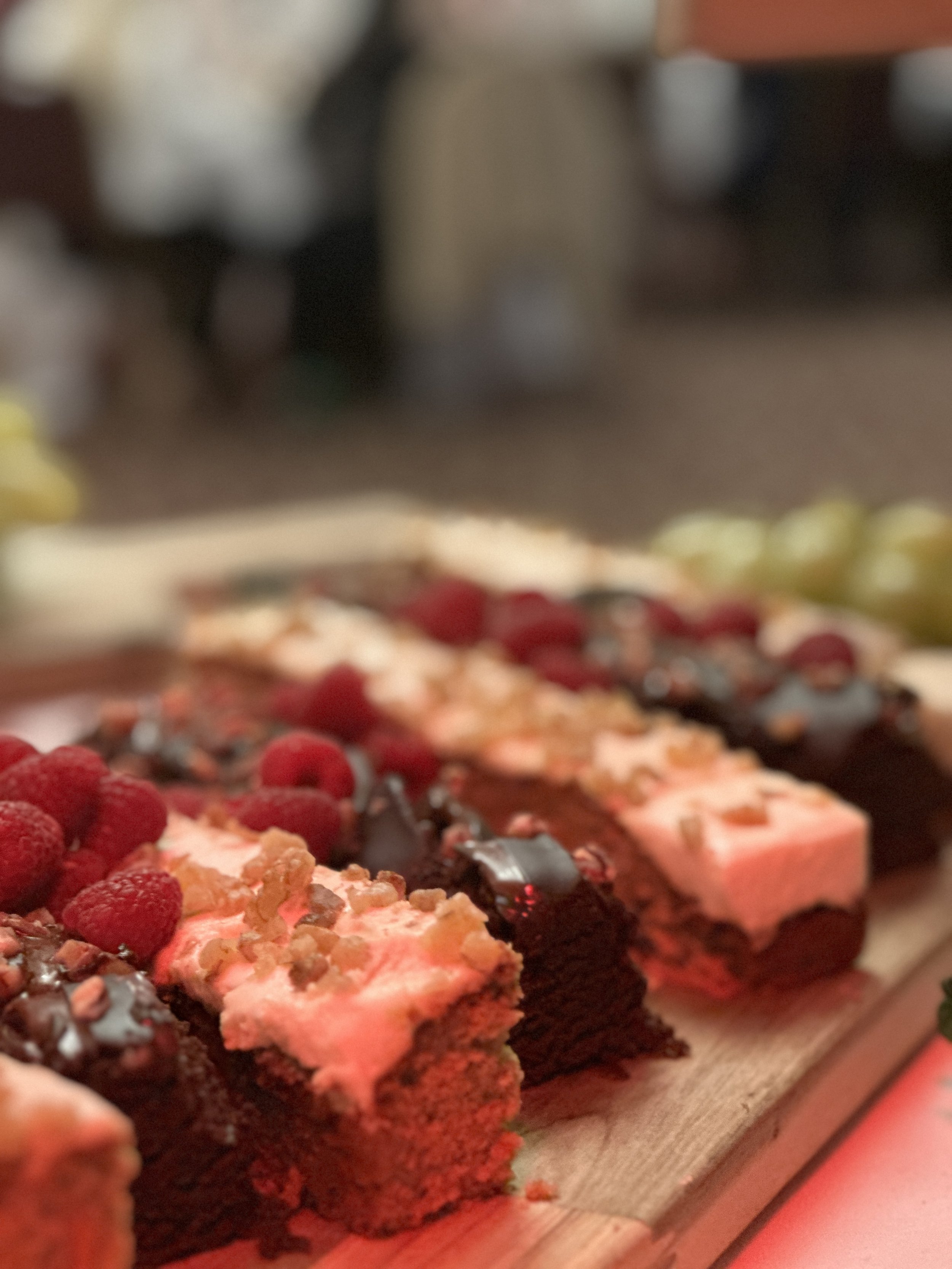 Close-up of assorted layered cakes topped with raspberries, chocolate drizzle, and nuts on a wooden board.