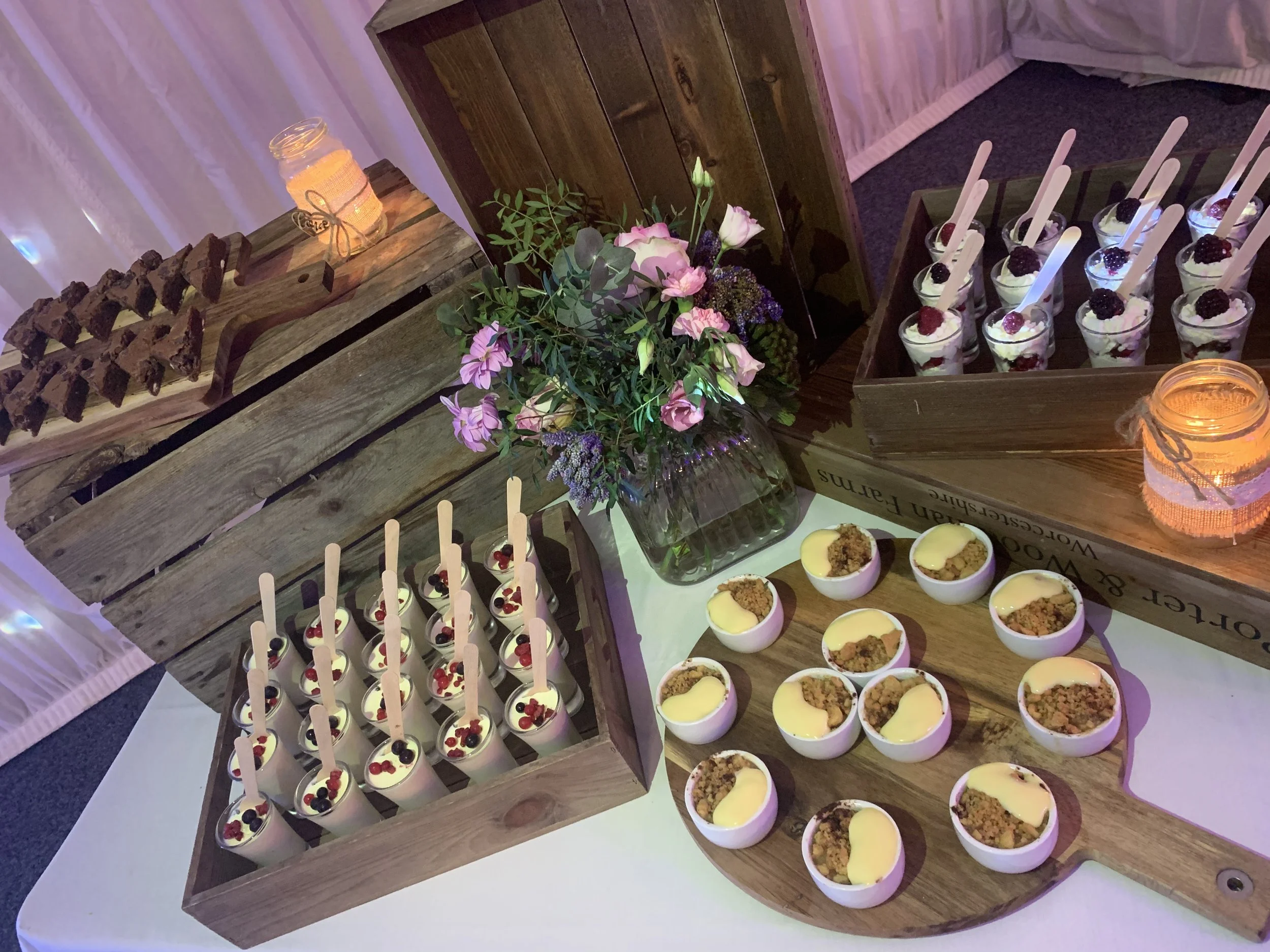 Dessert table with small cups of cheesecakes topped with fruit, arranged in wooden trays, with a floral centerpiece in a glass vase, and lit candles in jars.