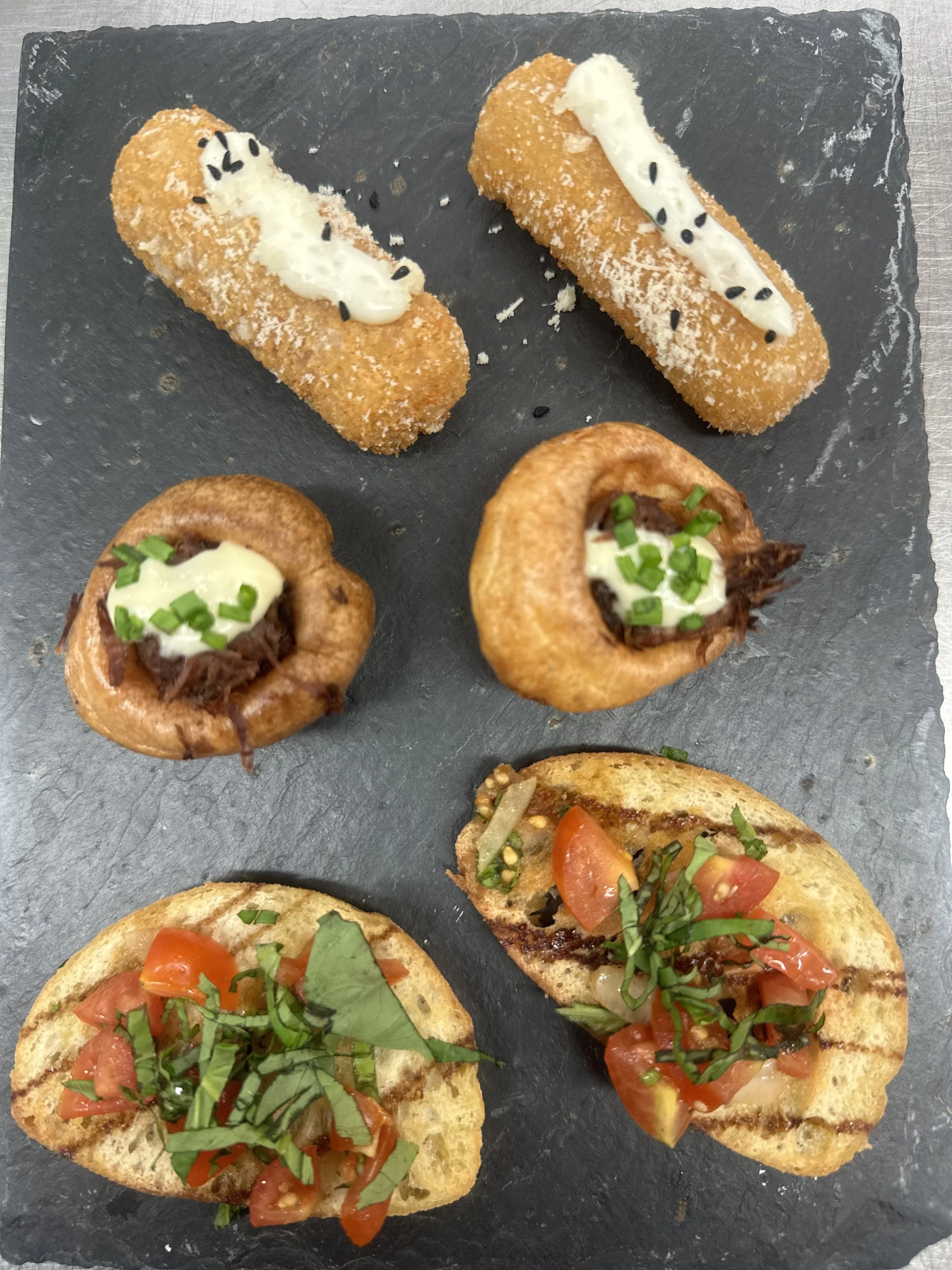 Assorted appetizers on a black slate platter, including bread topped with tomatoes and herbs, stuffed potato skins with cheese and green onions, and fried croquettes with sauces and black sesame seeds.