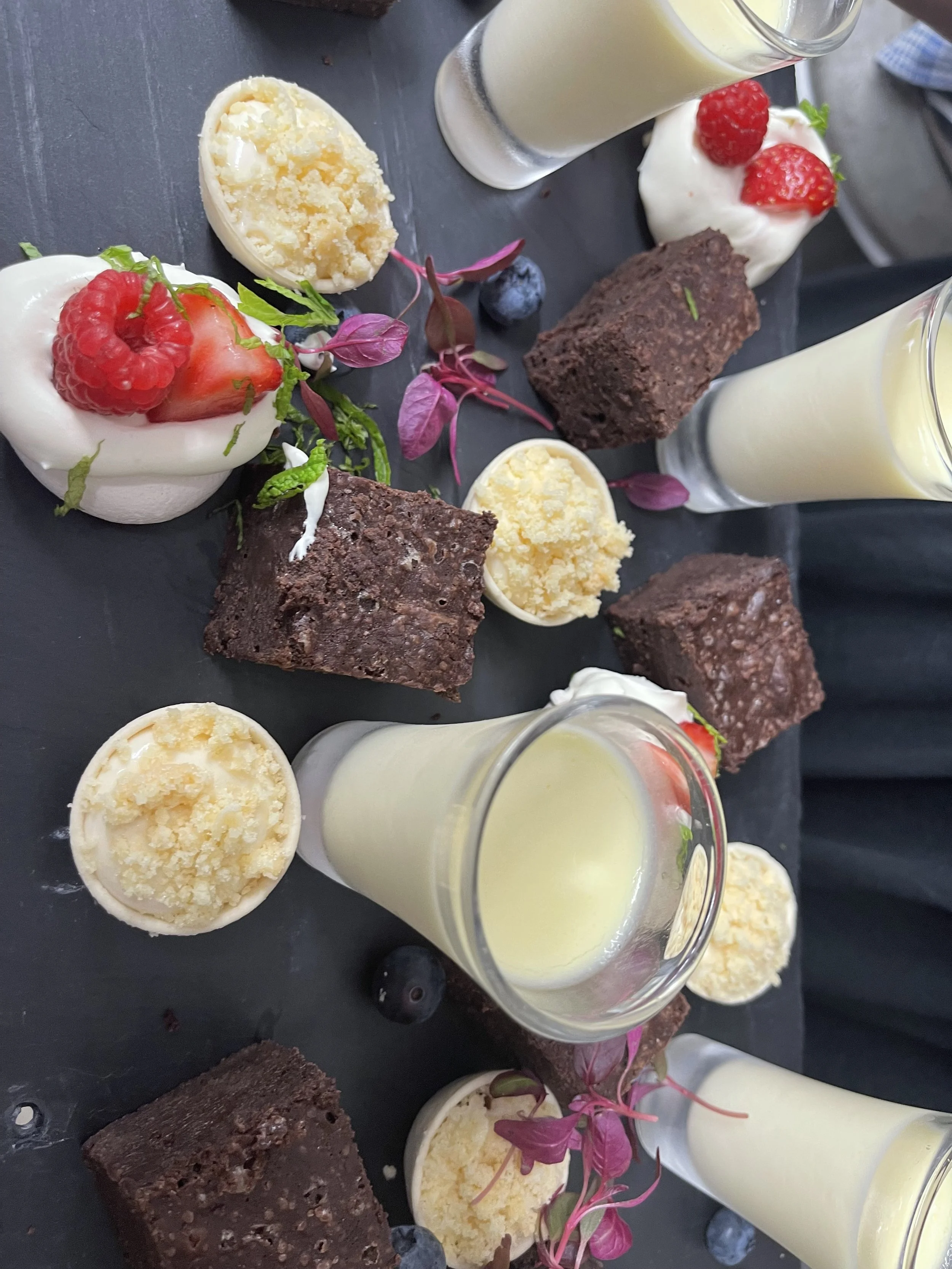 Dessert assortment includes glasses of vanilla pudding, mini chocolate brownies, vanilla cupcake topped with fresh strawberries and raspberries, and halved cupcakes with icing and fruit garnishes on a black platter.