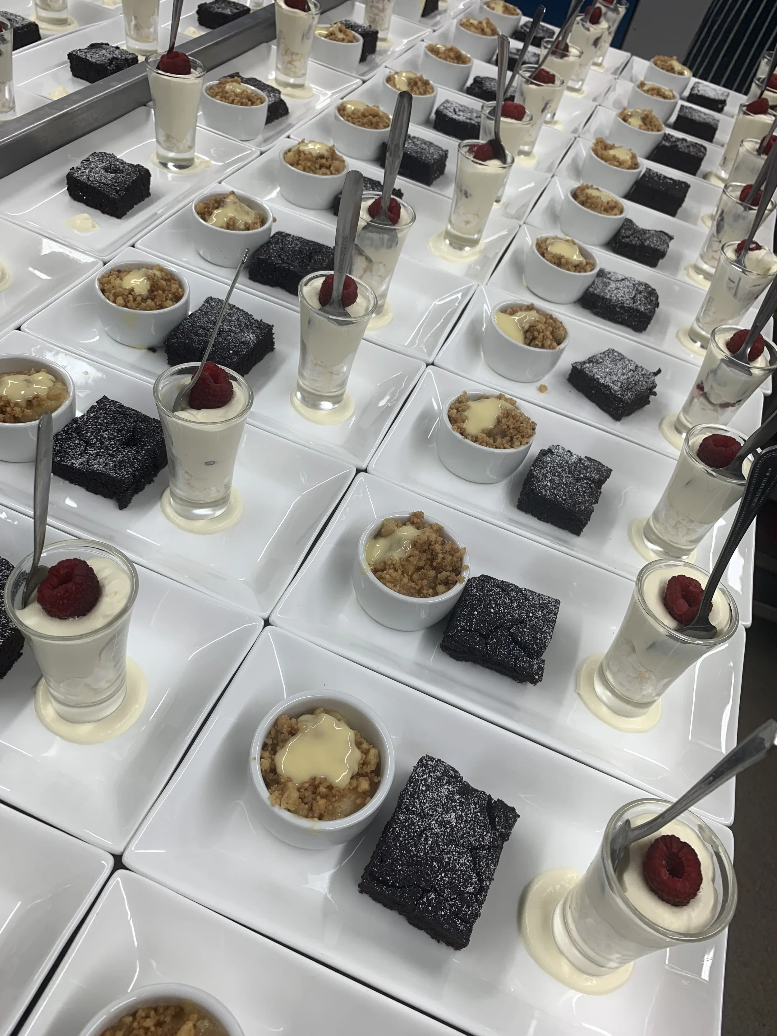 Multiple plates with chocolate brownies, mini cheesecakes topped with raspberries, and bowls of crumb-topped dessert with white sauce.