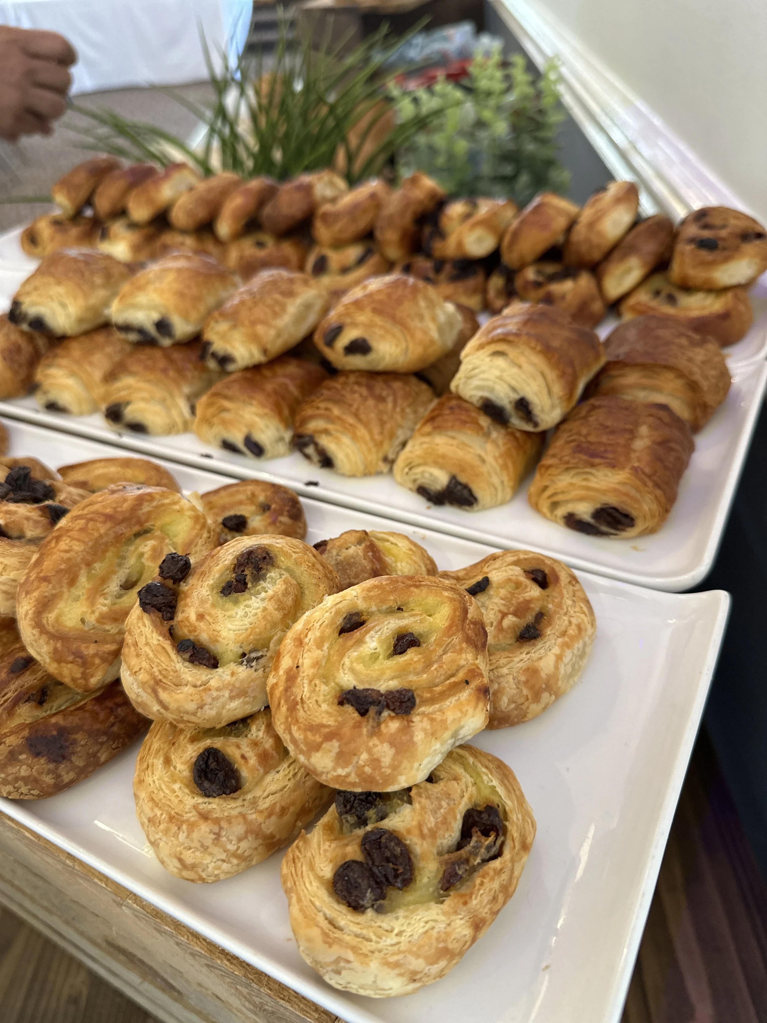 Assorted pastries on white platters, including chocolate croissants and Danish pastries with raisins.