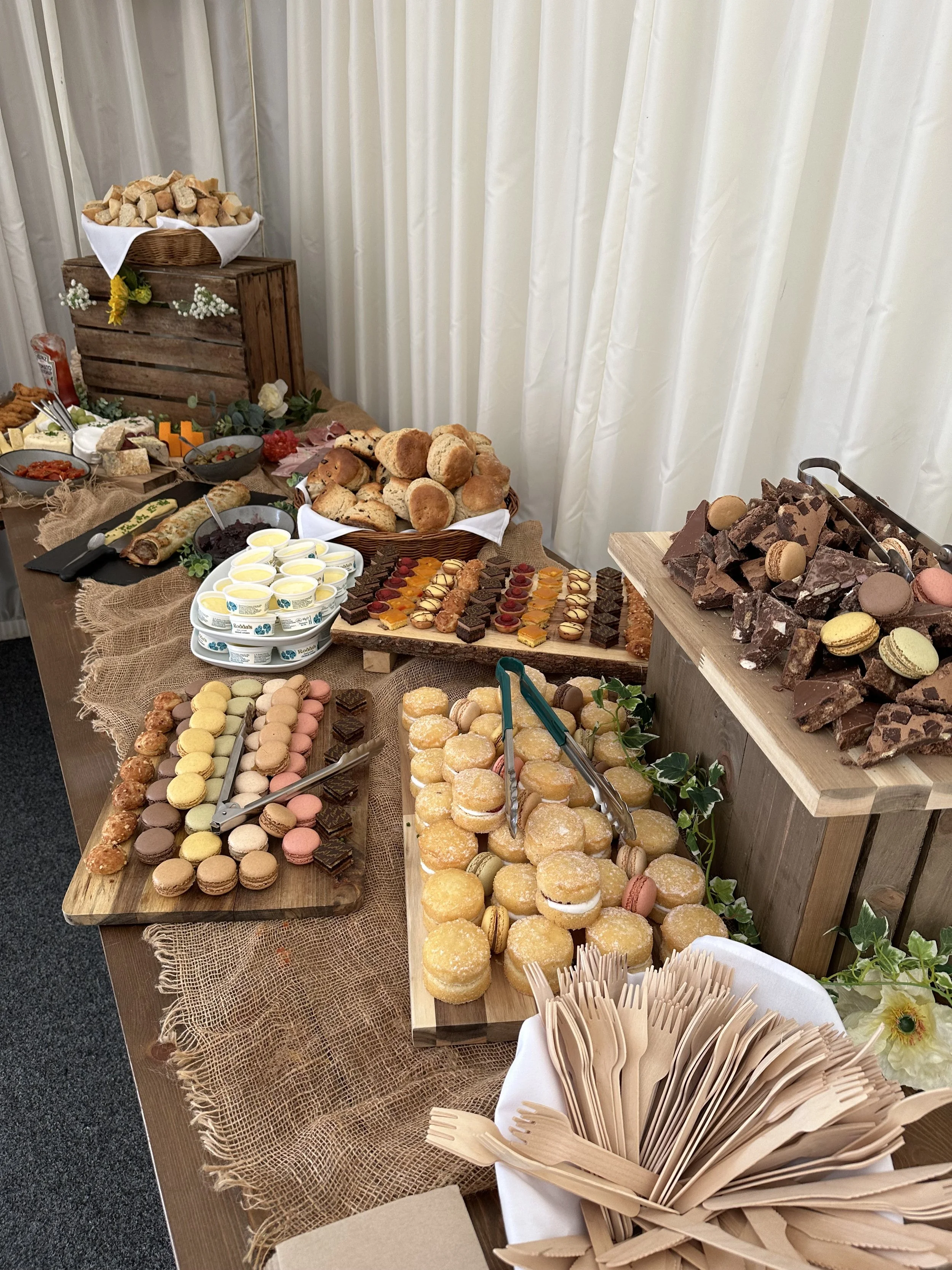 A buffet table with assorted bread, pastries, macarons, chocolates, and finger foods, decorated with burlap and greenery, set against white curtains.