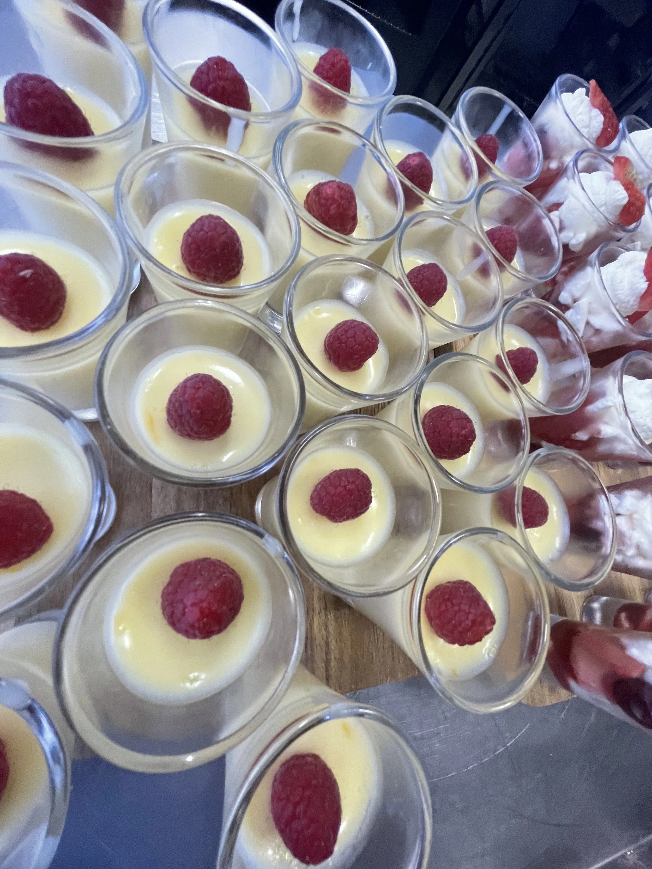 Several small glasses filled with a yellow dessert, topped with a raspberry, arranged on a wooden surface.