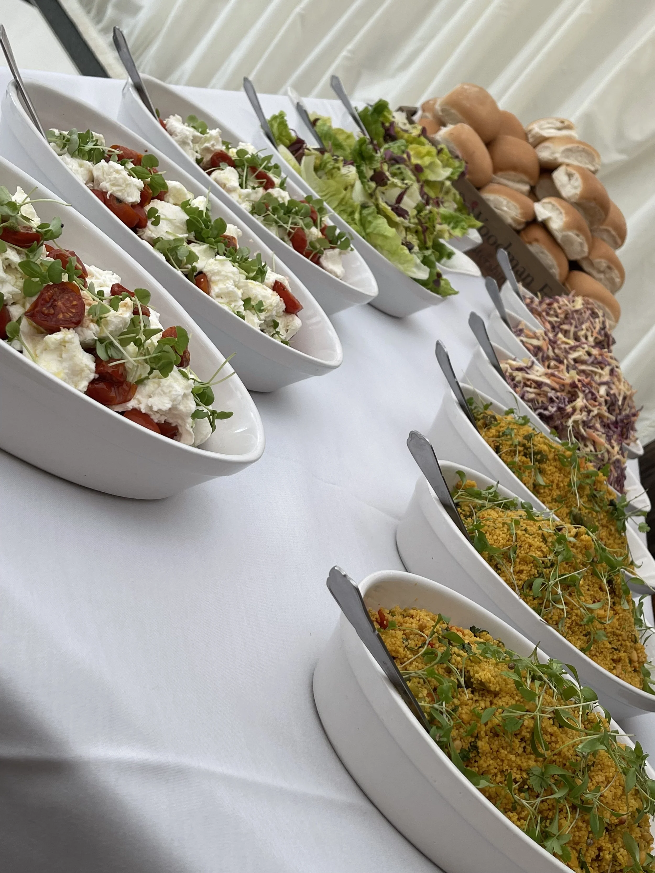 A variety of salads and bread rolls on a white tablecloth, including tomato and cheese salad, green salad, coleslaw, and mustard seed salad with microgreens.
