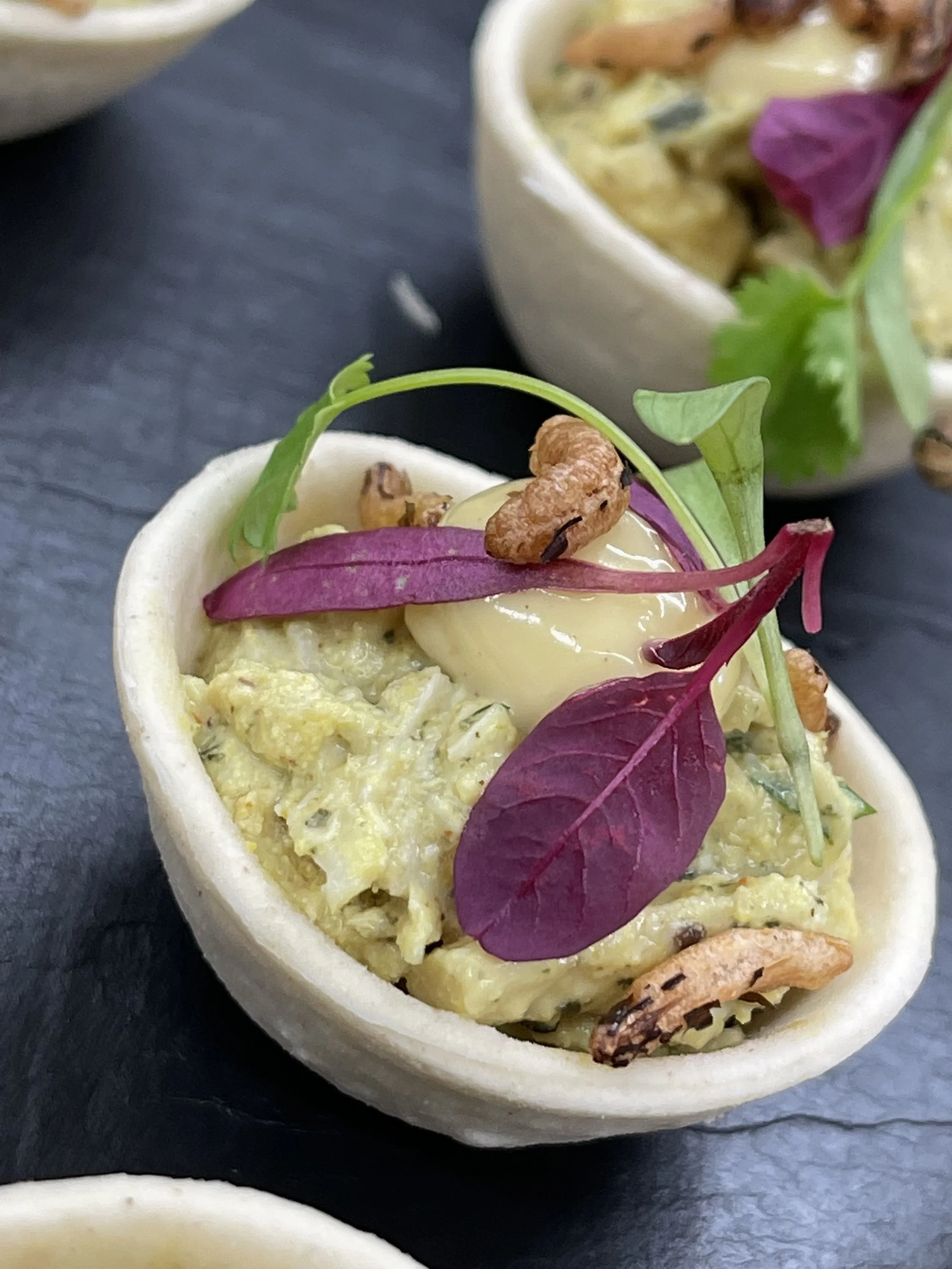 Close-up of a small tart filled with creamy avocado mixture, topped with microgreens, a quail egg, walnuts, and purple leaves.