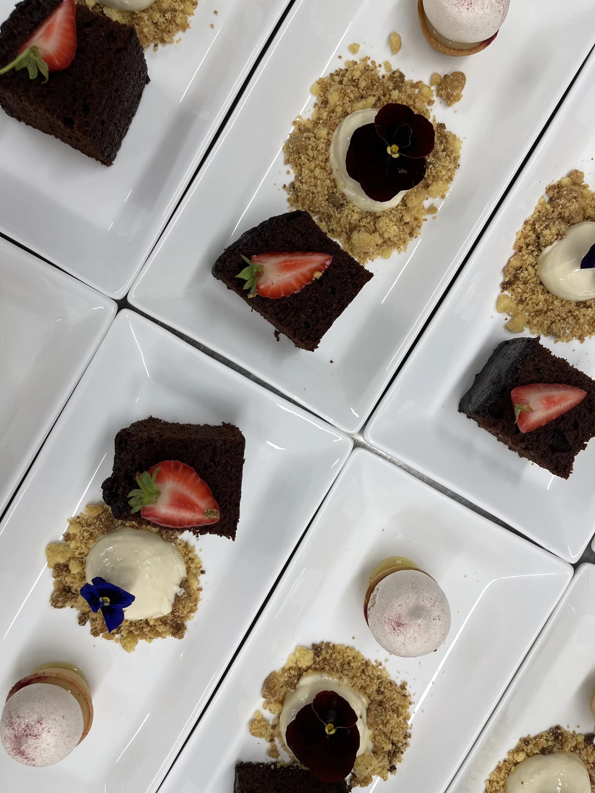 Multiple white rectangular plates with chocolate cake, strawberry garnish, crumbly toppings, cream, edible flowers, and macarons.