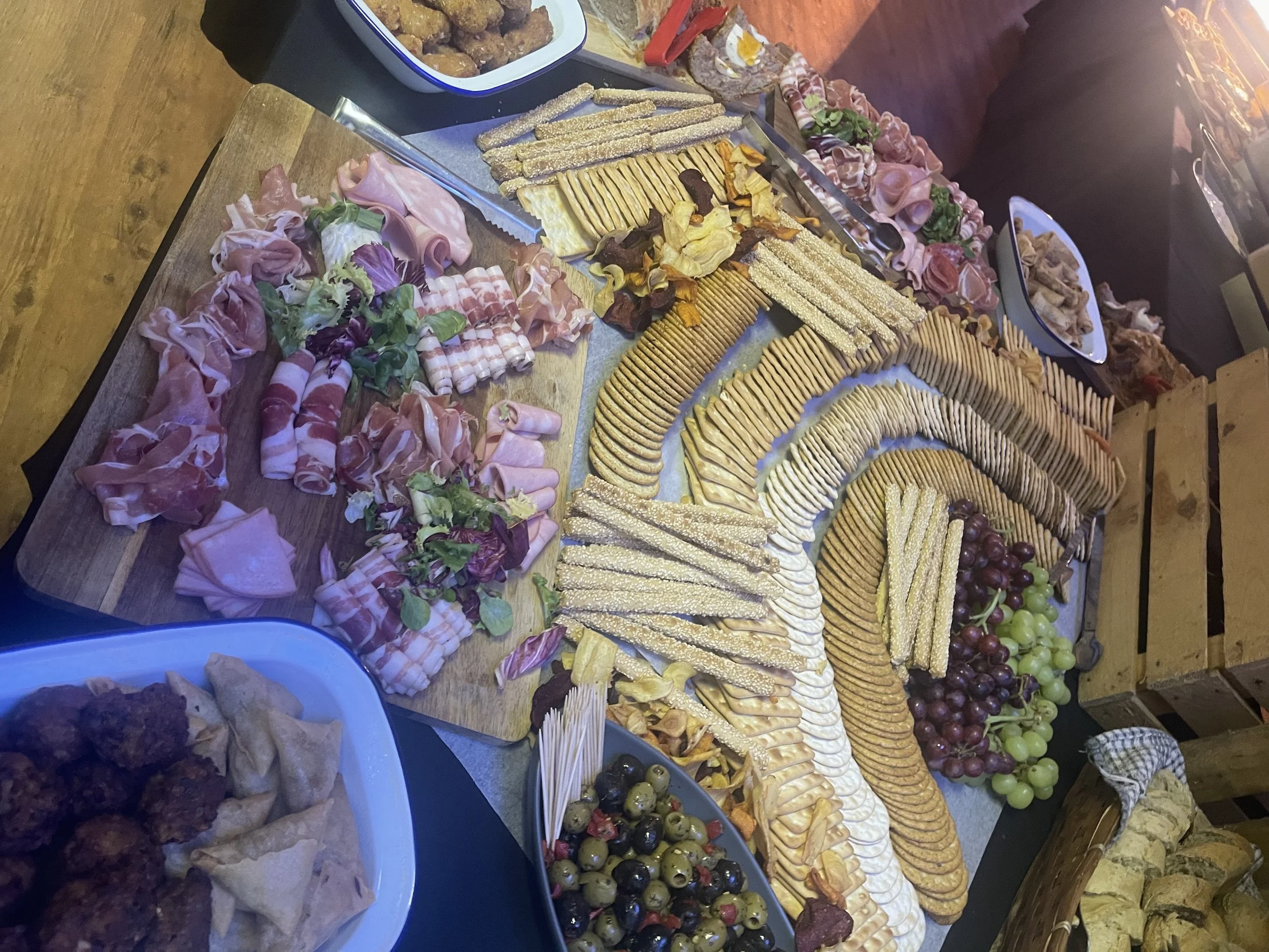 A cheese and charcuterie platter with various types of sliced cheeses, cured meats, grapes, olives, and bread, arranged on a large wooden board.