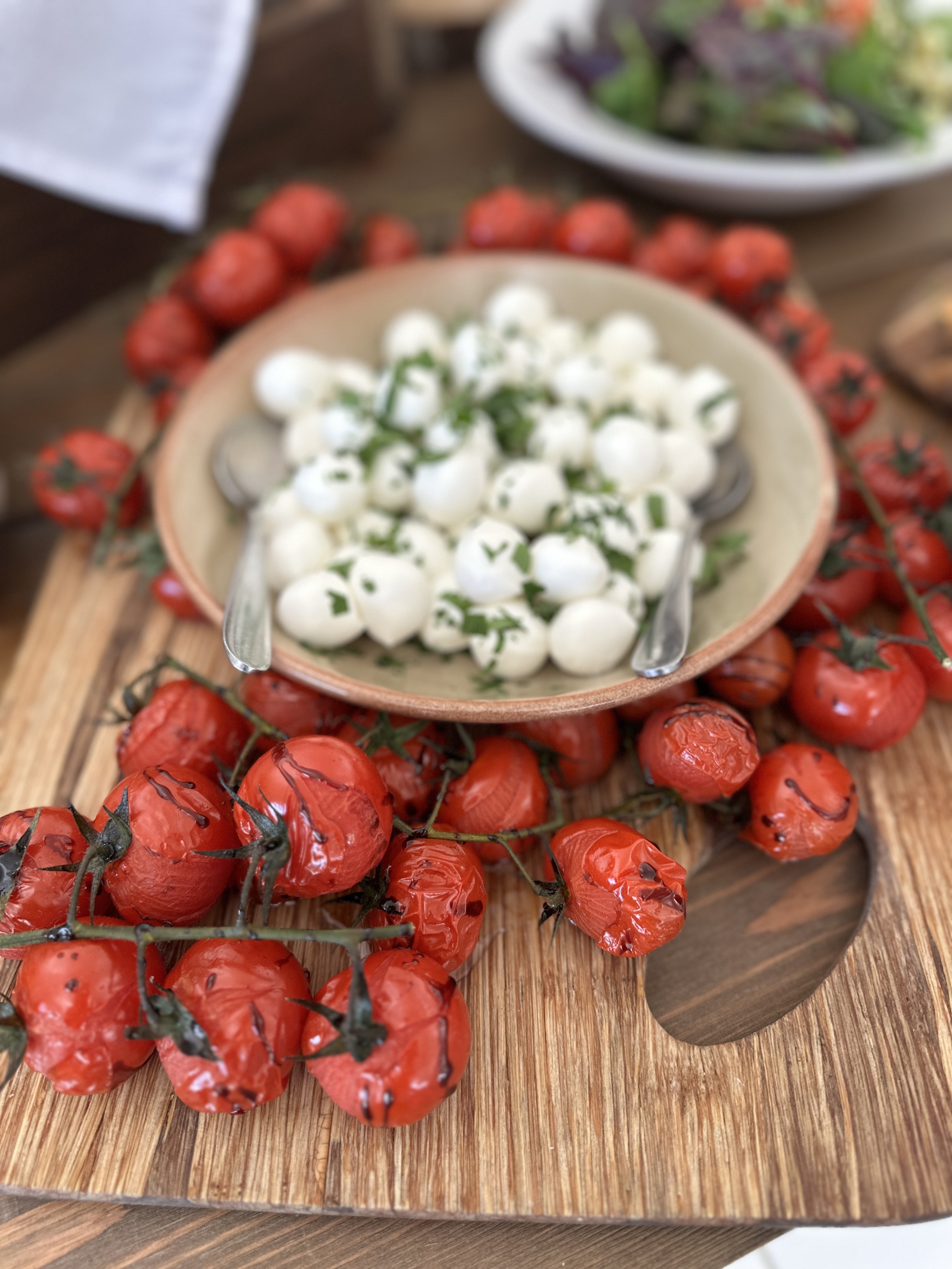 A bowl of small white mozzarella balls garnished with herbs on a wooden serving board surrounded by roasted cherry tomatoes with balsamic glaze.
