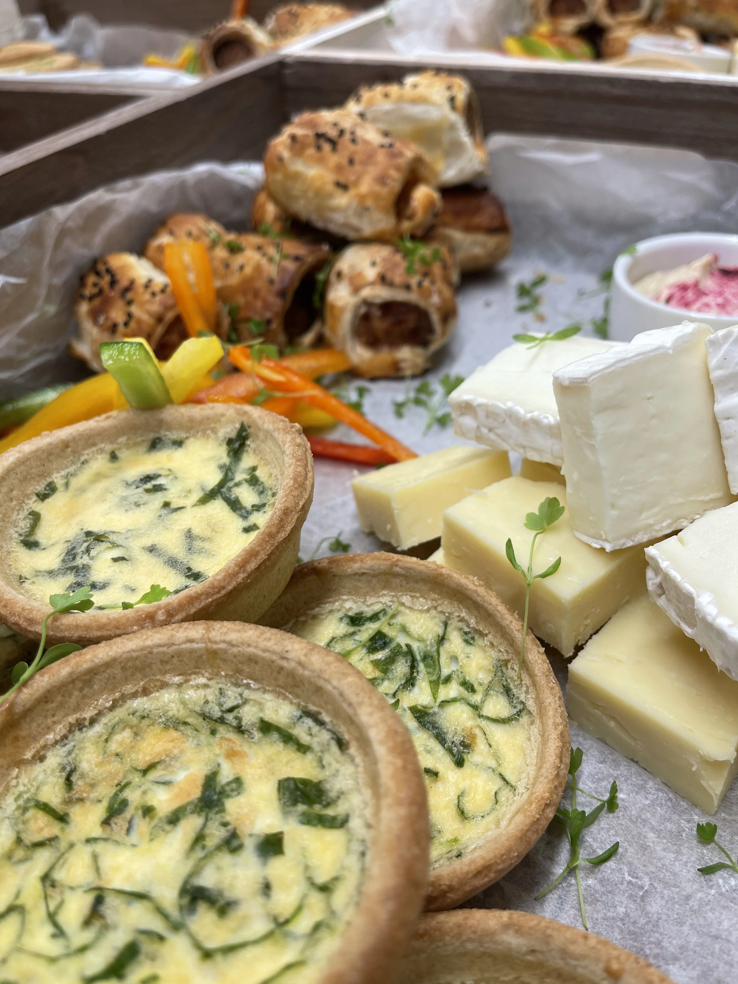 A tray of assorted savory snacks including mini quiches with spinach, sliced cheeses, and vegetable rolls garnished with herbs.