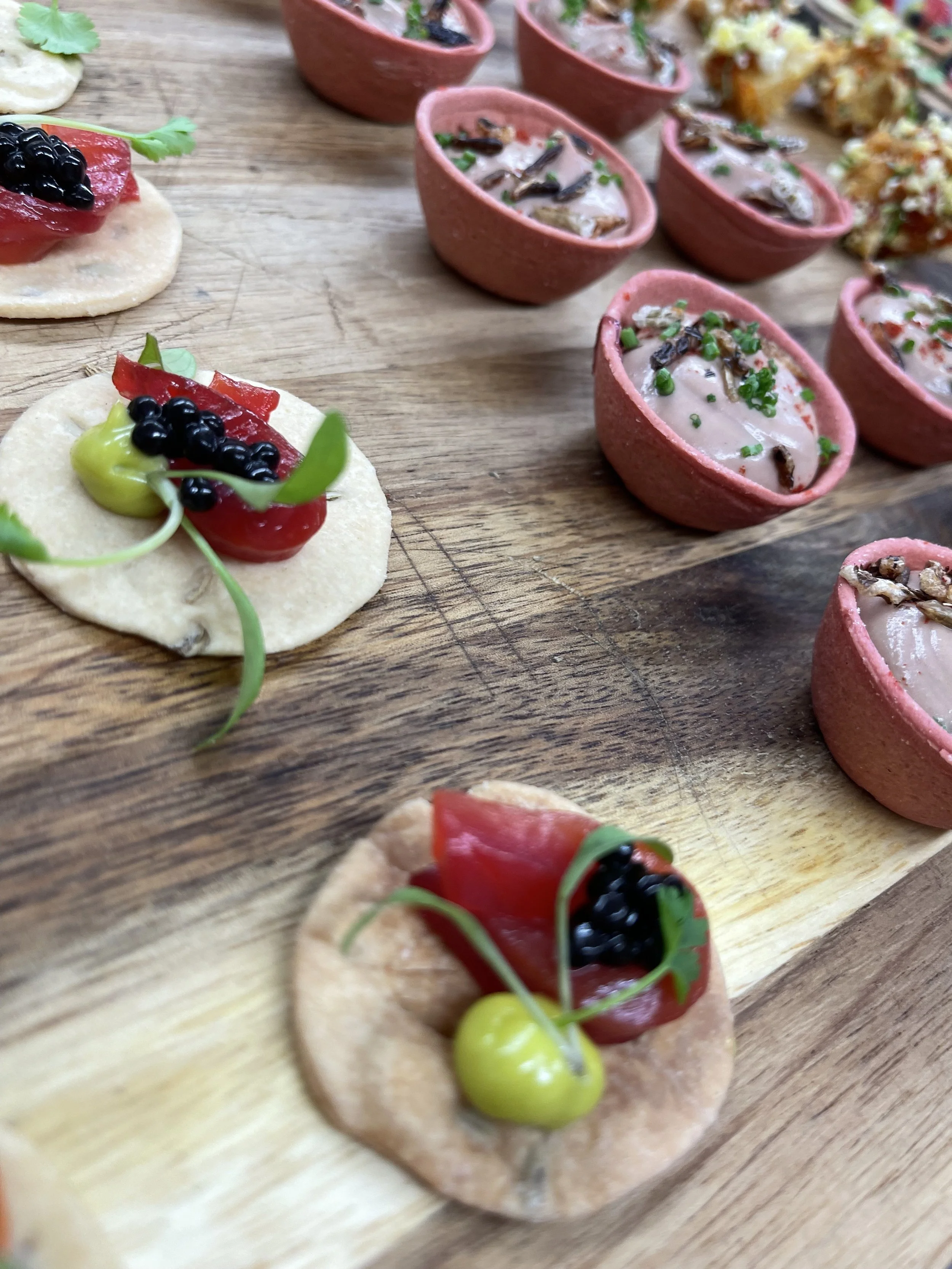 Close-up of assorted appetizers on a wooden board: tiny crackers topped with red and green toppings, and small pink bowls filled with creamy dip garnished with herbs and small fish.