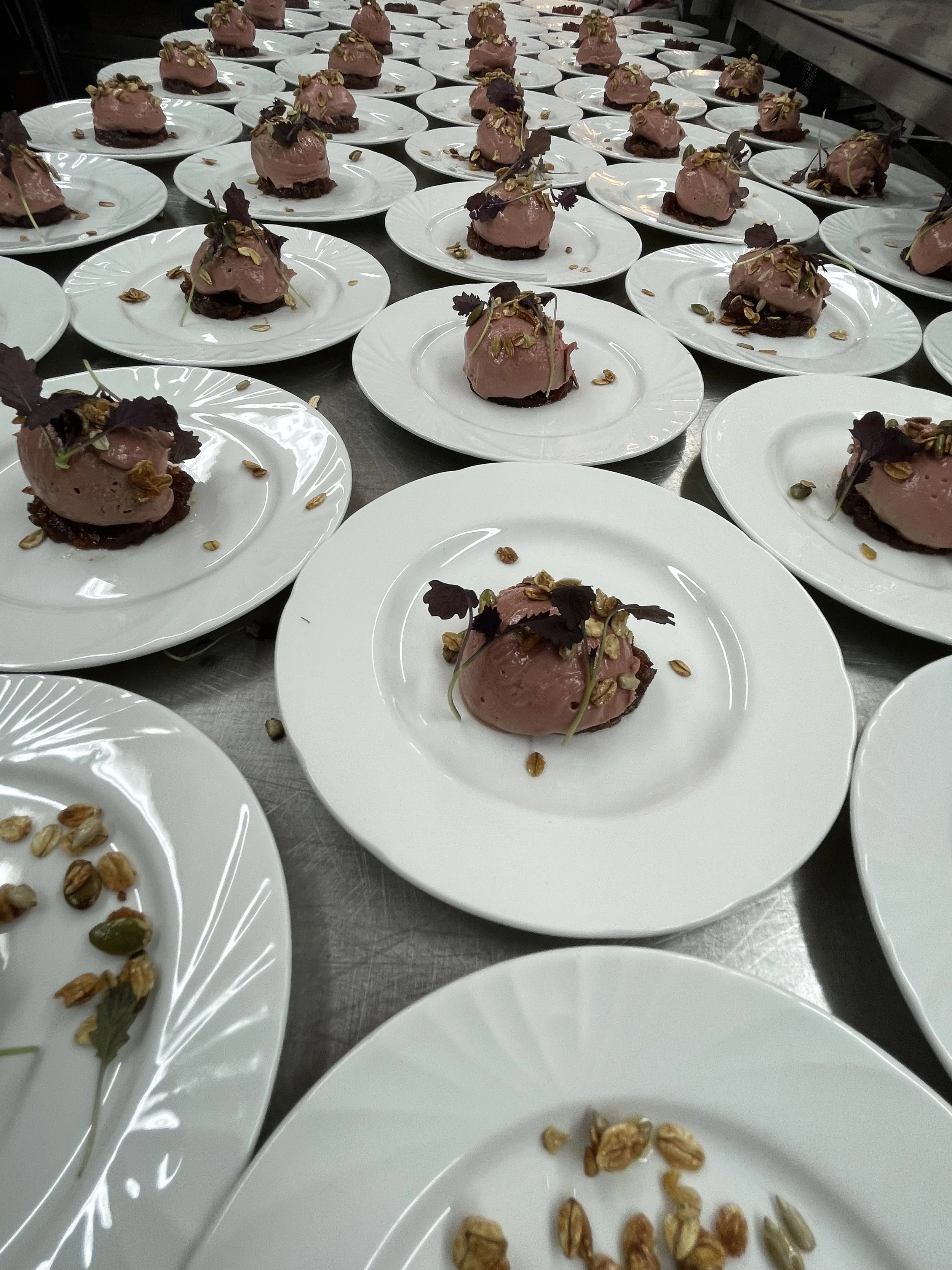 Multiple plates with pink mousse desserts garnished with chocolate pieces, nuts, and leaves, arranged on a stainless steel table.