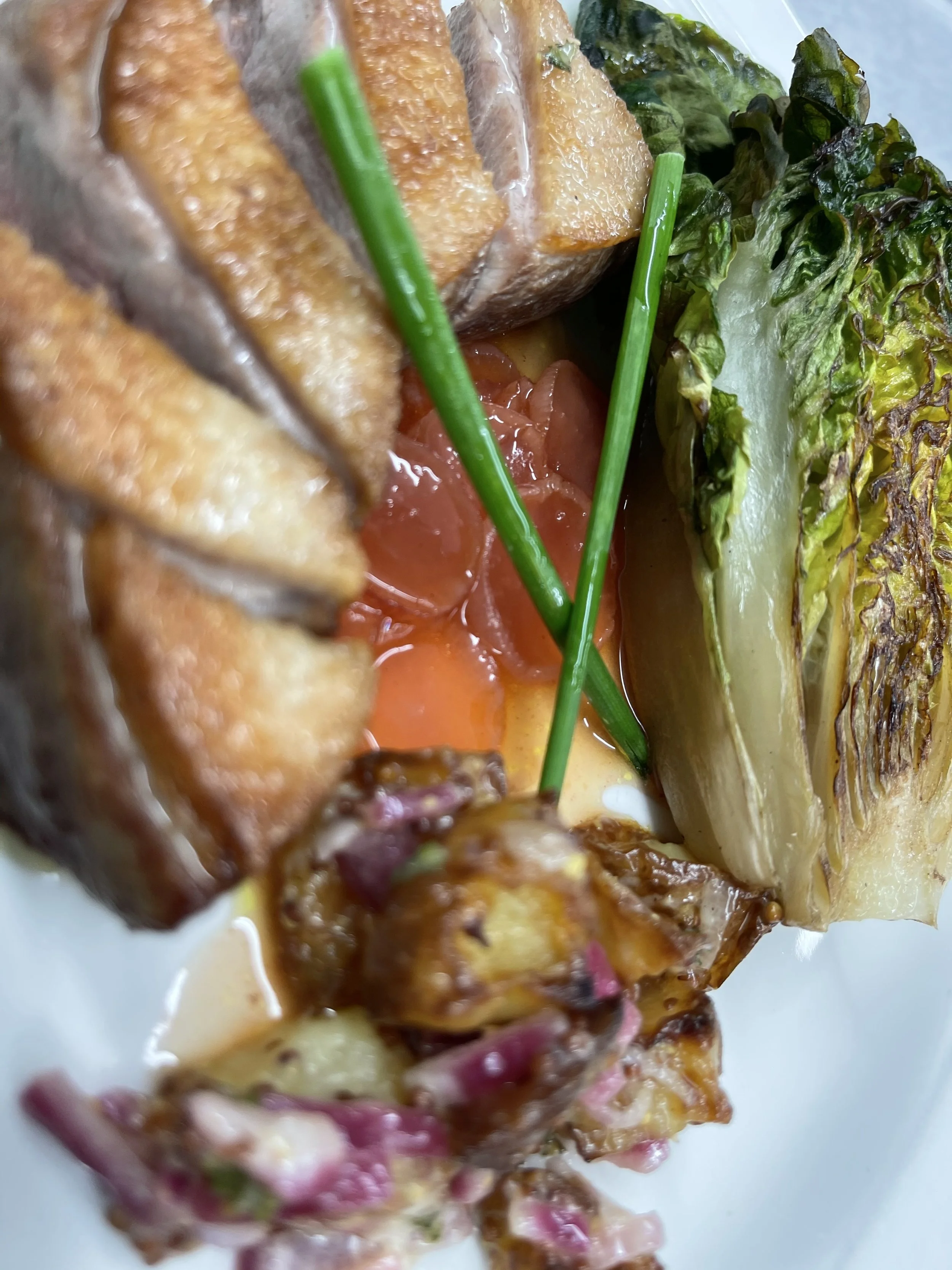 Close-up of a gourmet dish featuring fried fish chunks, cherry tomatoes, grilled lettuce, grilled onion, and green chives.