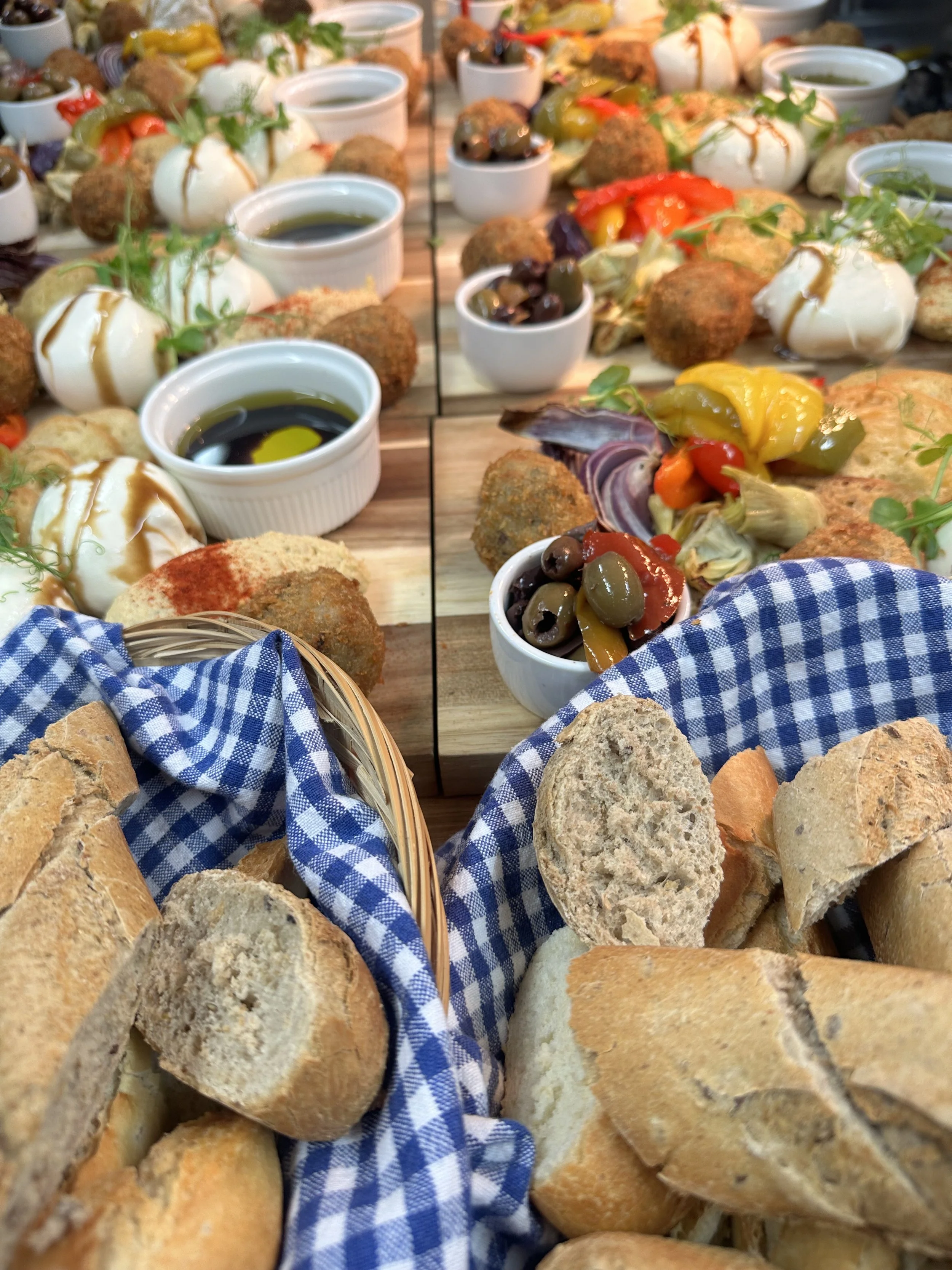 A table with baskets of bread, plates of assorted Mediterranean appetizers, and small bowls of olive oil and balsamic vinegar, set for a meal.