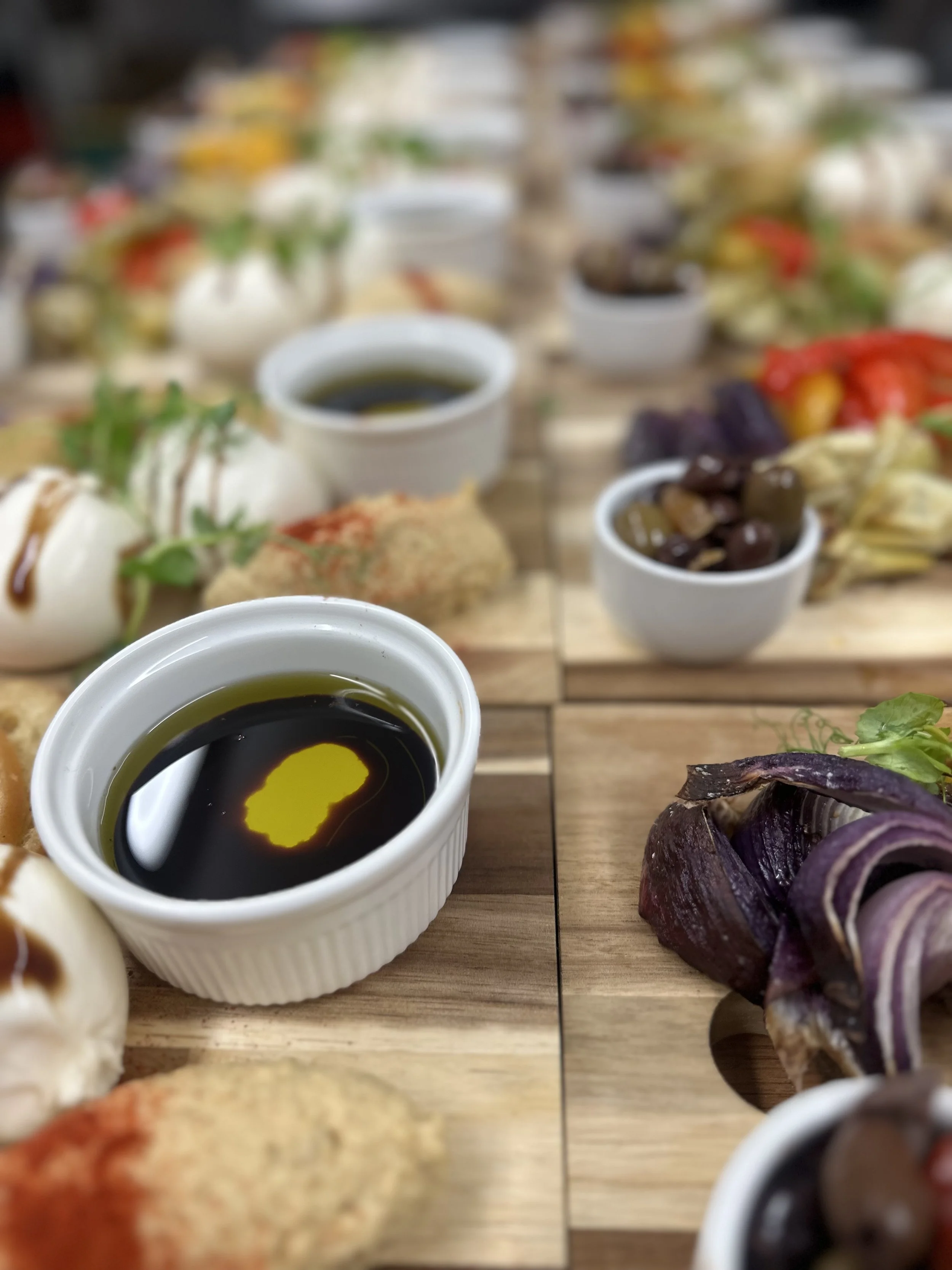 A close-up of various appetizers and dips on a wooden platter, including a small white ramekin of soy sauce, roasted vegetables, and a mozzarella ball with basil, on a table with more dishes in the background.