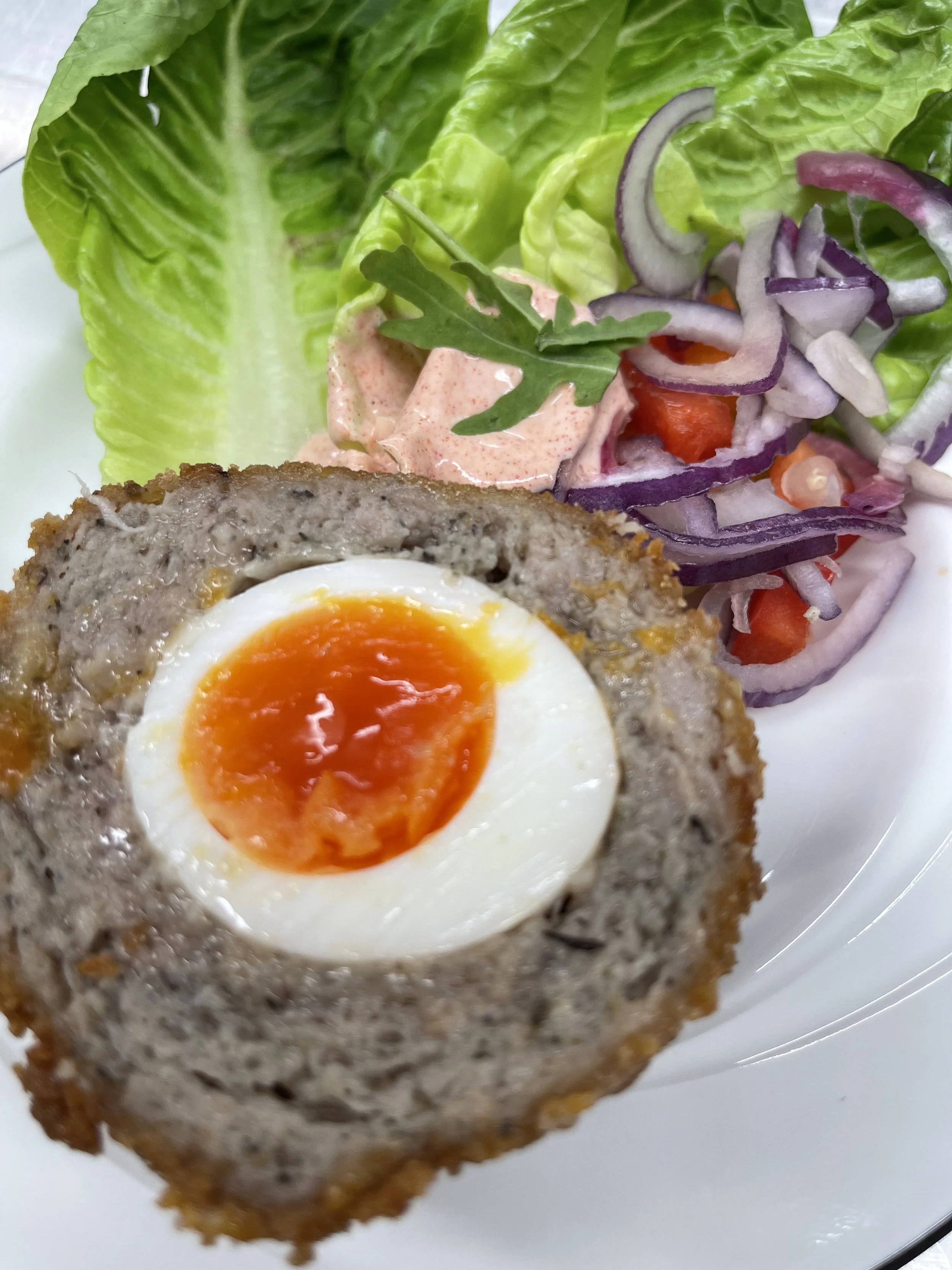 Close-up of a Scotch egg with a runny yolk, served with salad greens, sliced onions, tomatoes, and a creamy sauce.