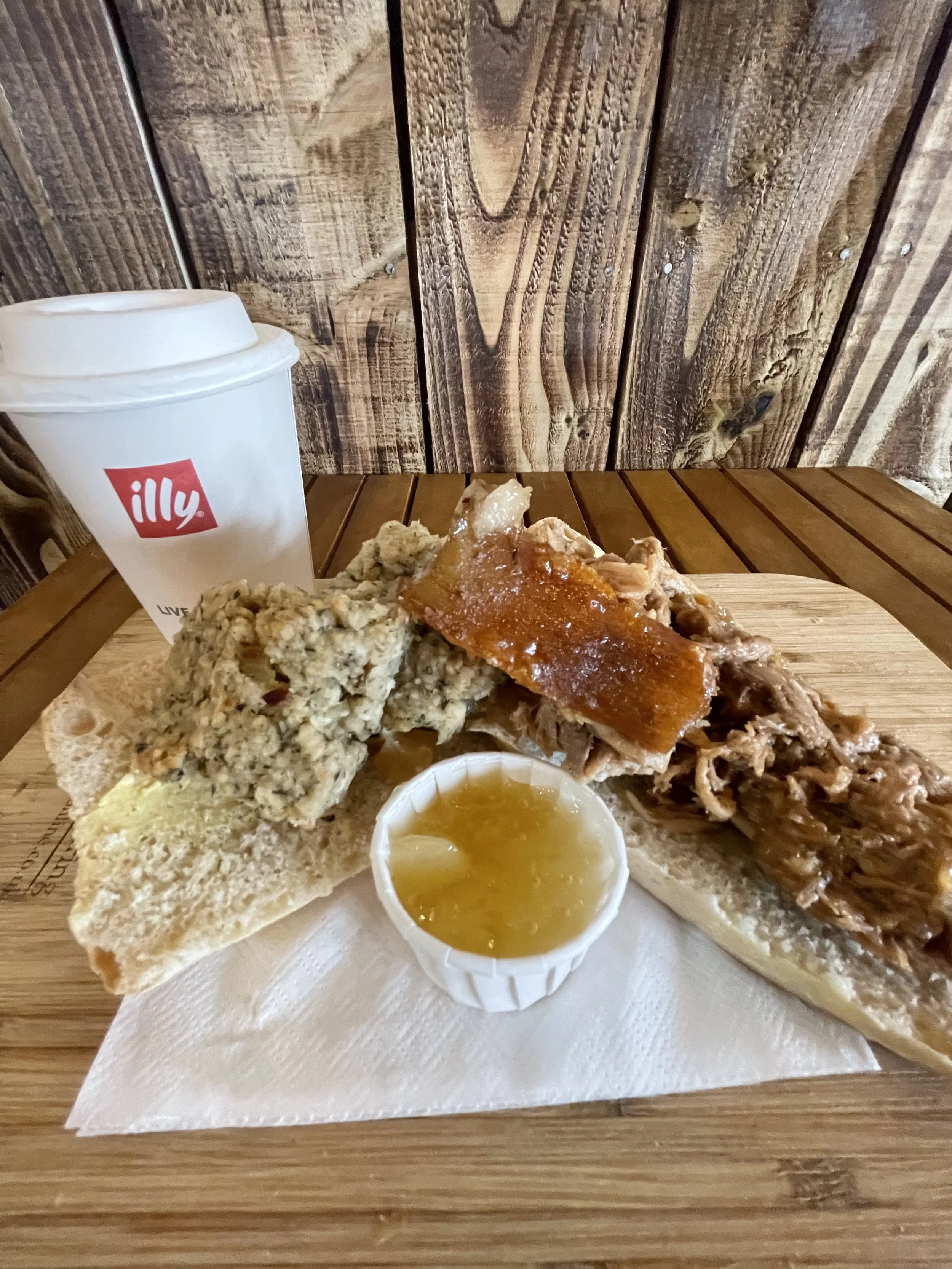 Sandwich platter with bread, assorted meats, a small cup of gravy or sauce, a soft drink, and a side of biscuit, on a wooden plate.
