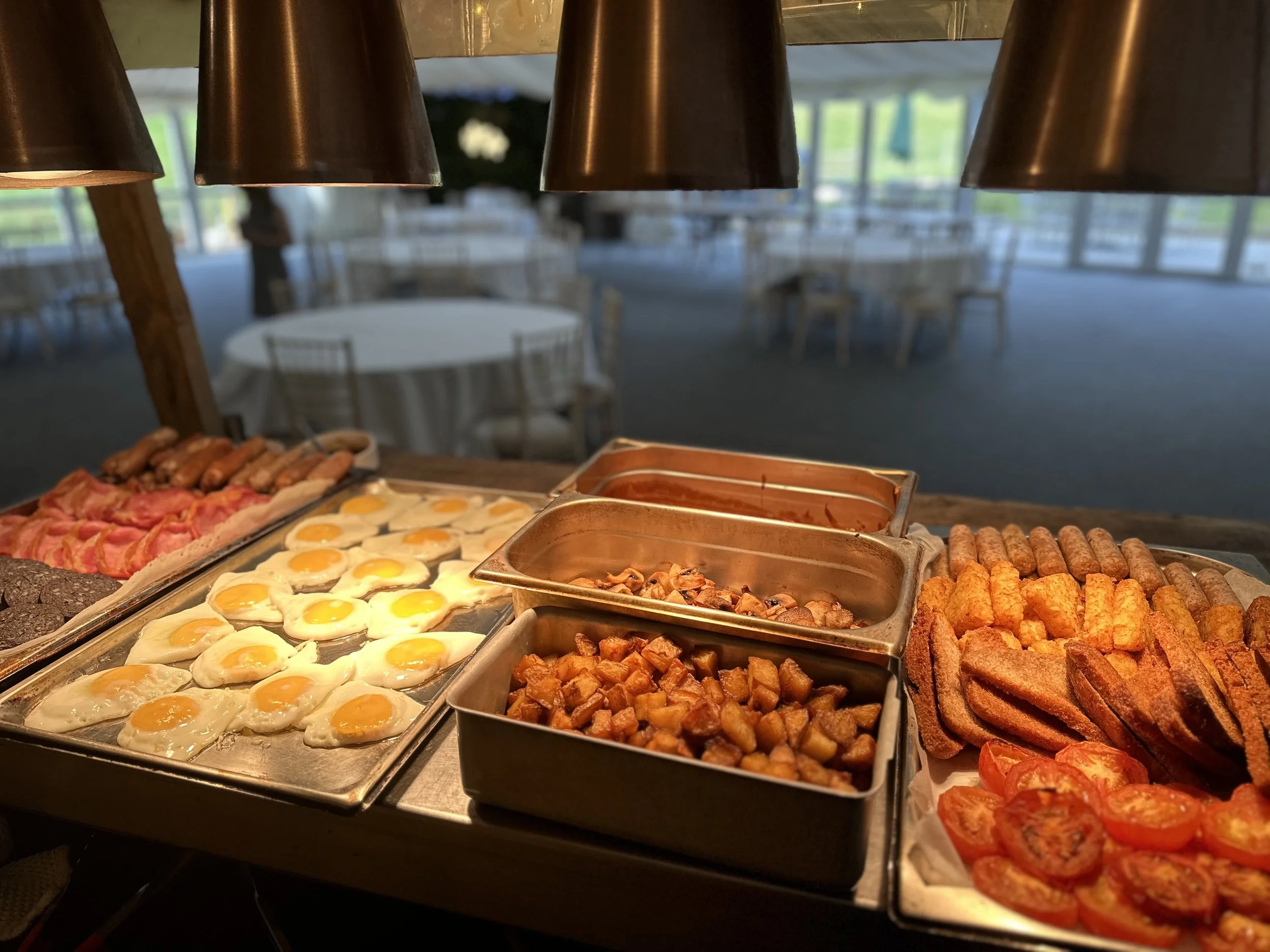 Breakfast buffet with fried eggs, bacon, sausages, hash browns, and toast, set up in a dining area with empty tables and chairs in the background.