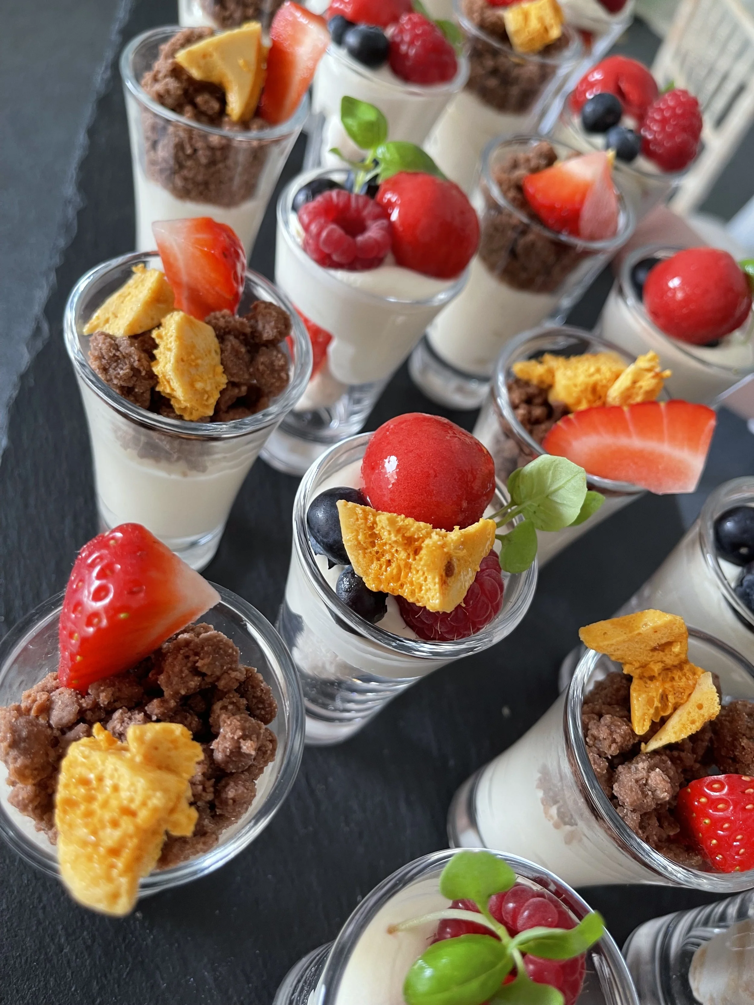 Assorted fruit parfaits topped with strawberries, blueberries, raspberries, and garnished with basil leaves, served in small glasses.