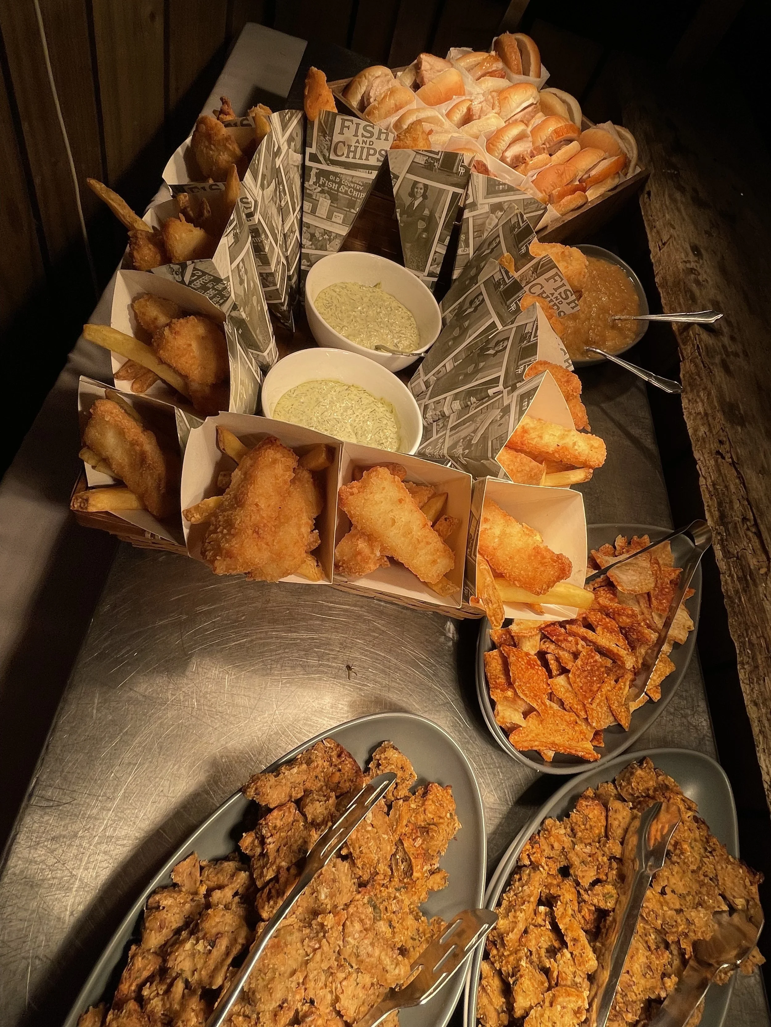 A variety of fried seafood, fish sticks, and chips served in small paper boxes, along with bowls of tartar sauce and a bowl of orange-colored seafood sauce, arranged on a wooden table.