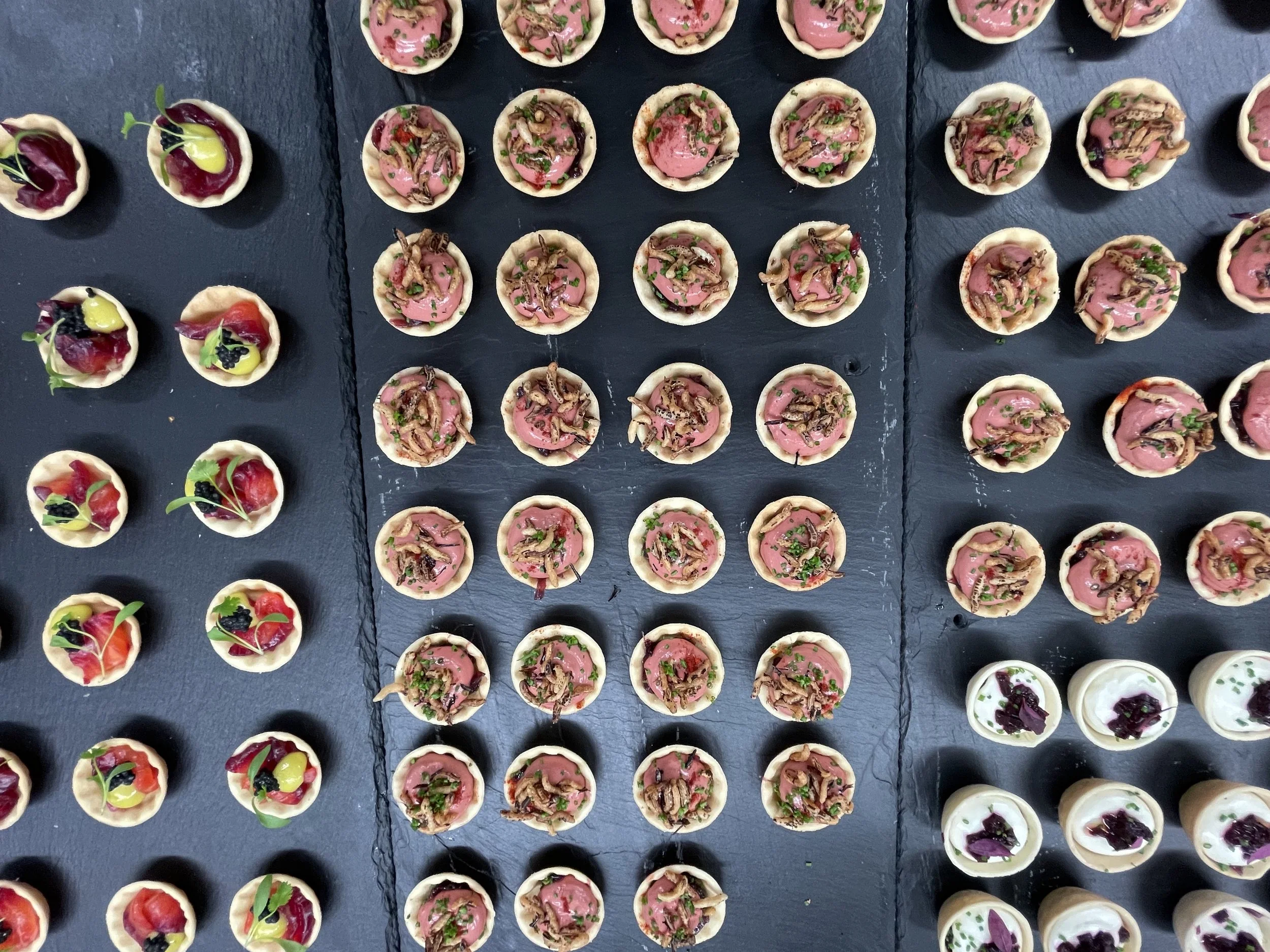 Various small appetizer tartlets arranged on black trays. The tartlets contain different toppings including pinkish filling with herbs and some with red and green garnishes, and others with white filling topped with purple leaves.